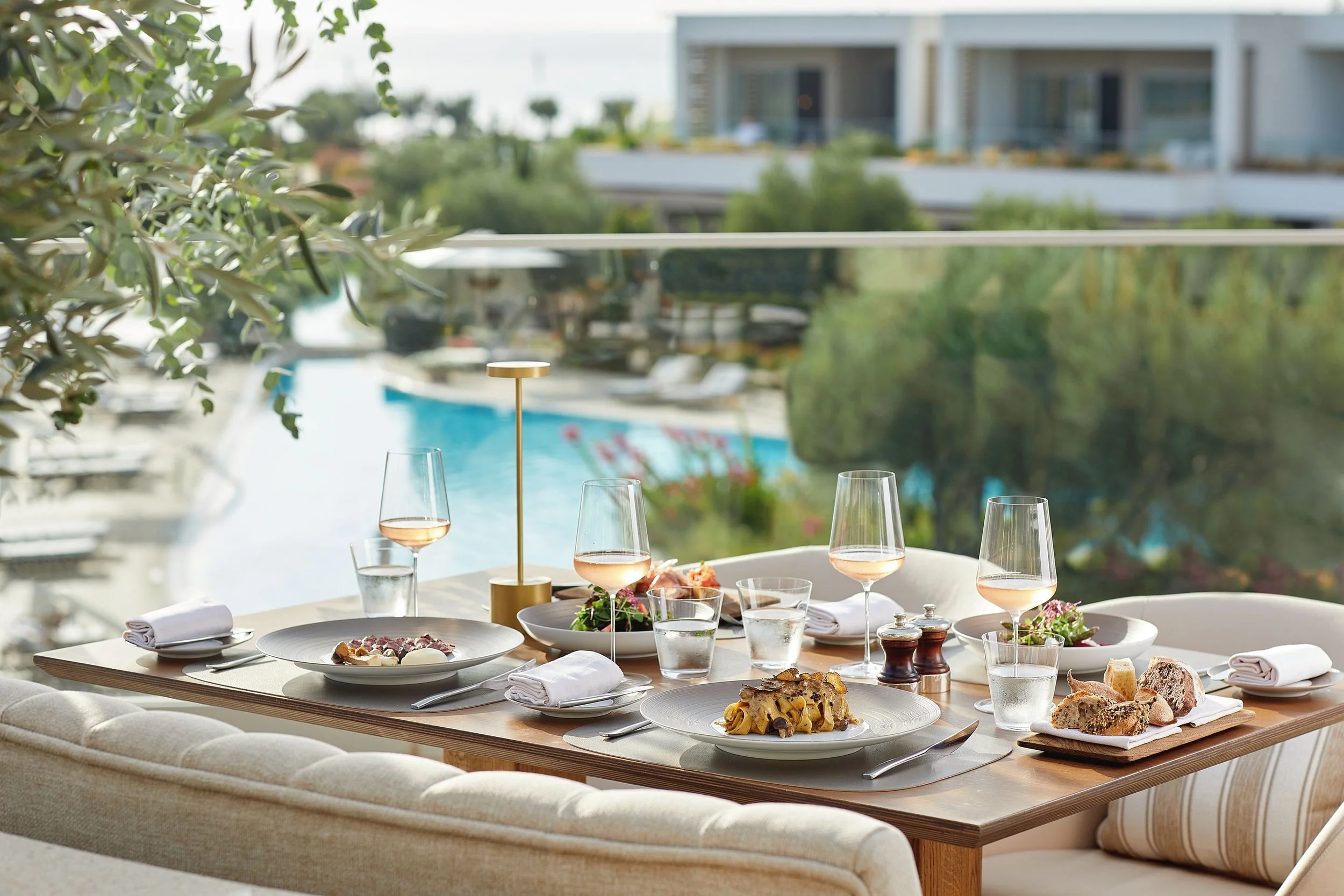 A table set for dining with plates of food, wine glasses, cutlery, and napkins, near a window with a view of a pool, trees, and a modern white building.