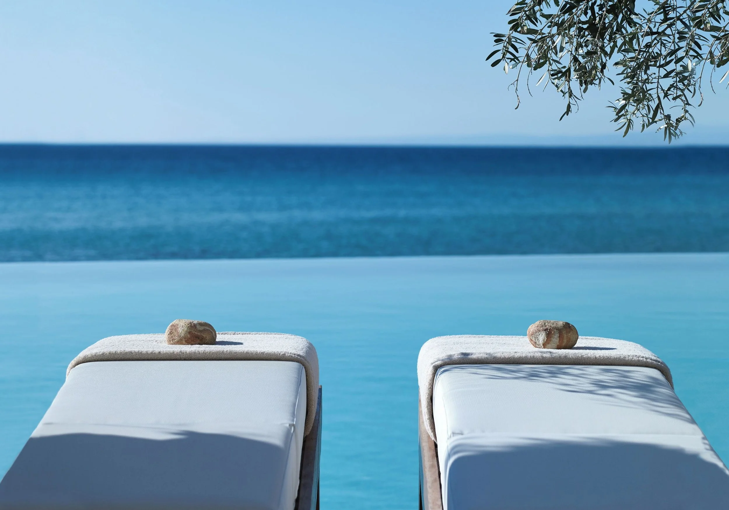 Two white lounge chairs with rocks on top, facing the ocean on a sunny day with a clear blue sky, and tree branches in the top right corner.