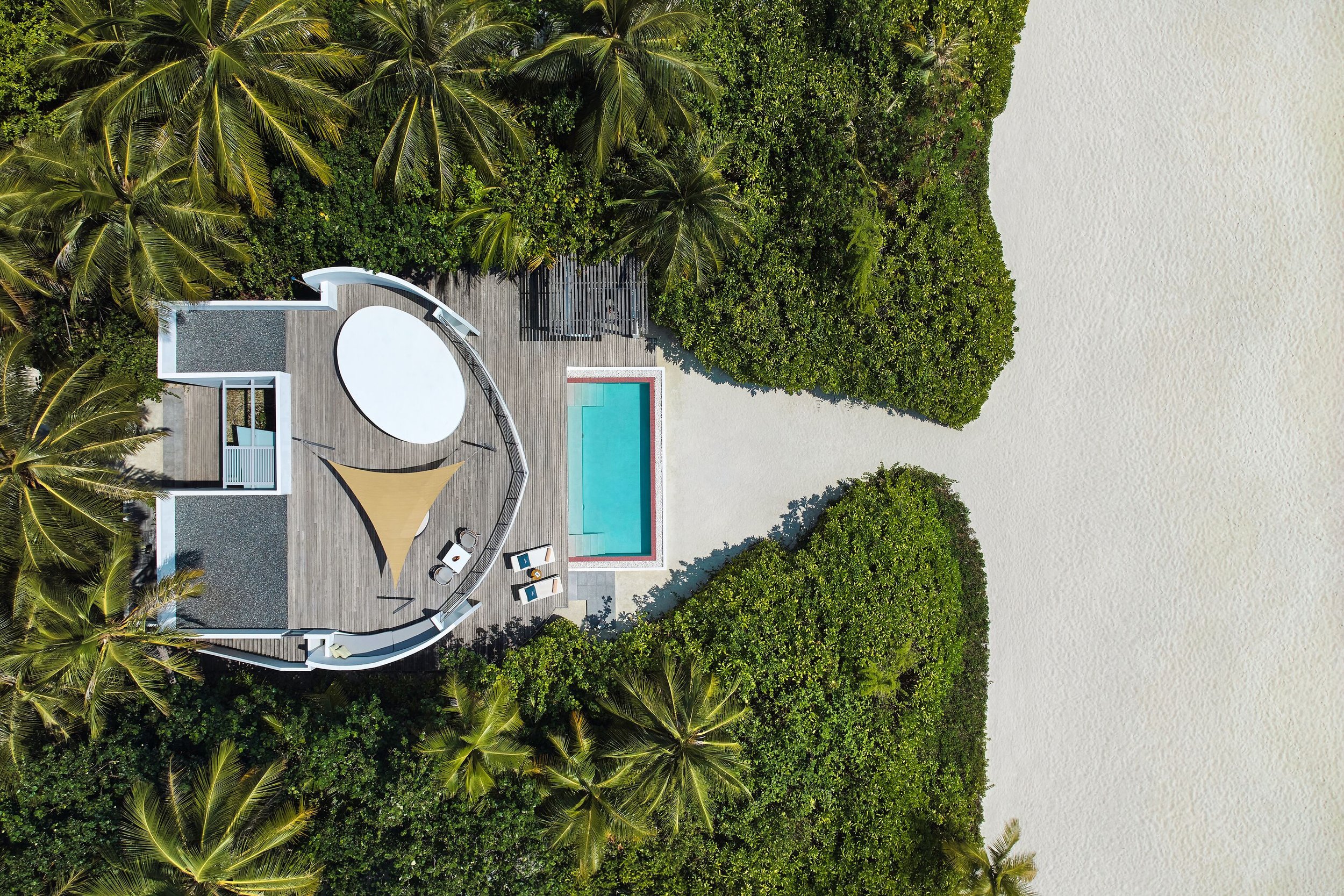 Aerial view of a tropical resort with a wooden deck, a swimming pool, lounge chairs, and surrounding lush green palm trees and shrubs, overlooking the ocean.