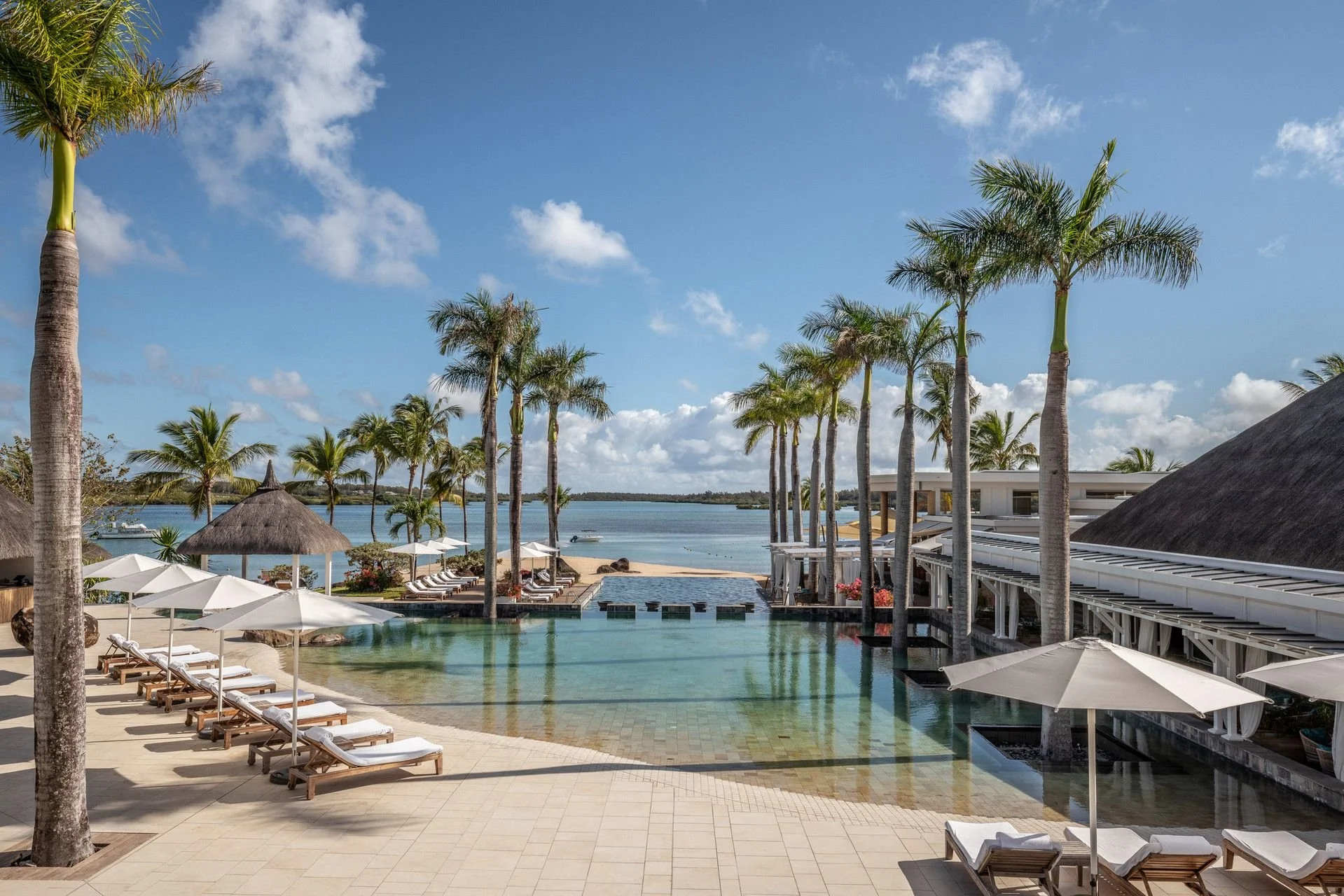 Beachside resort with palm trees, pool area with lounge chairs and umbrellas, and a view of the ocean under a partly cloudy sky.