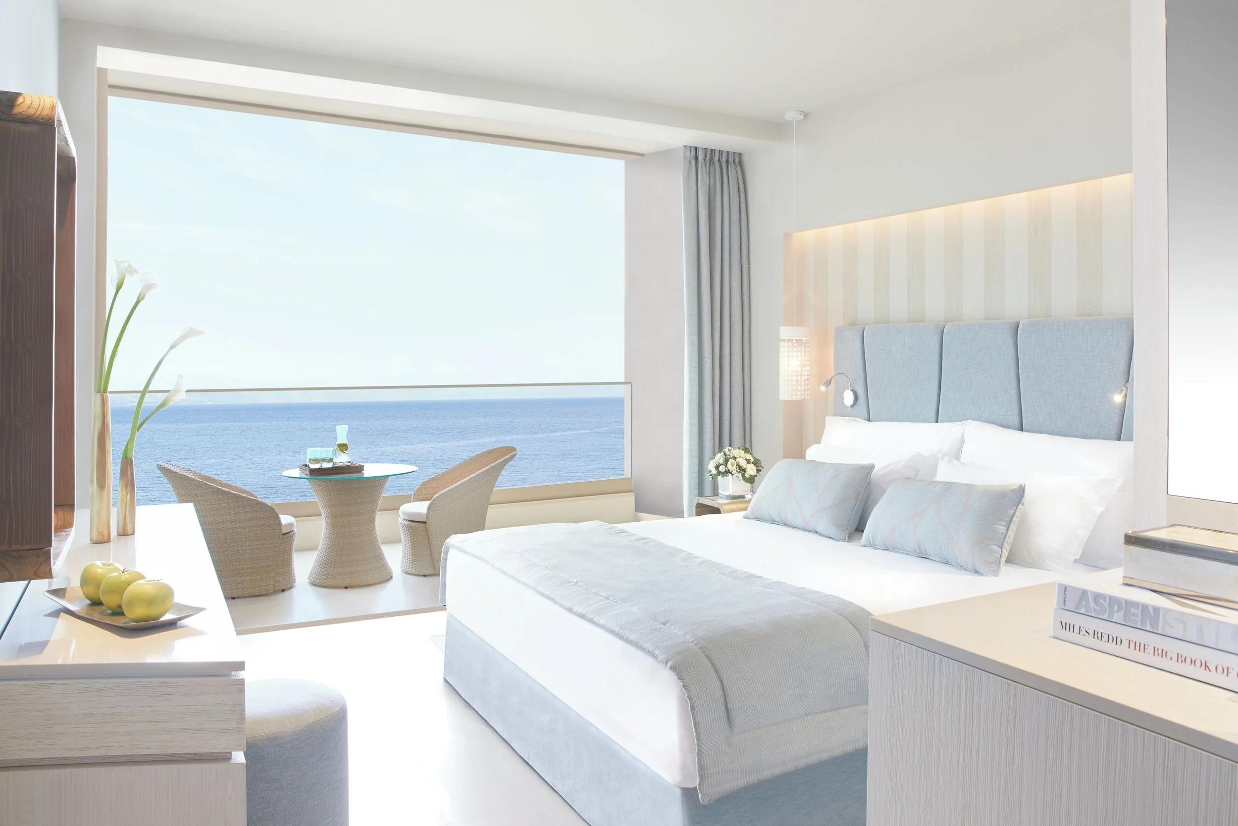 A modern hotel room with a large window showing an ocean view, a bed with a light blue headboard, a sitting area with a round table and two chairs, and decorative plants and lamps.