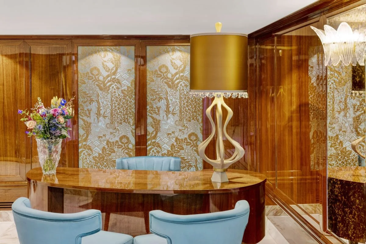 Elegant office with a wooden desk, blue armchairs, a floral arrangement, a tall table lamp, and decorative wallpaper.