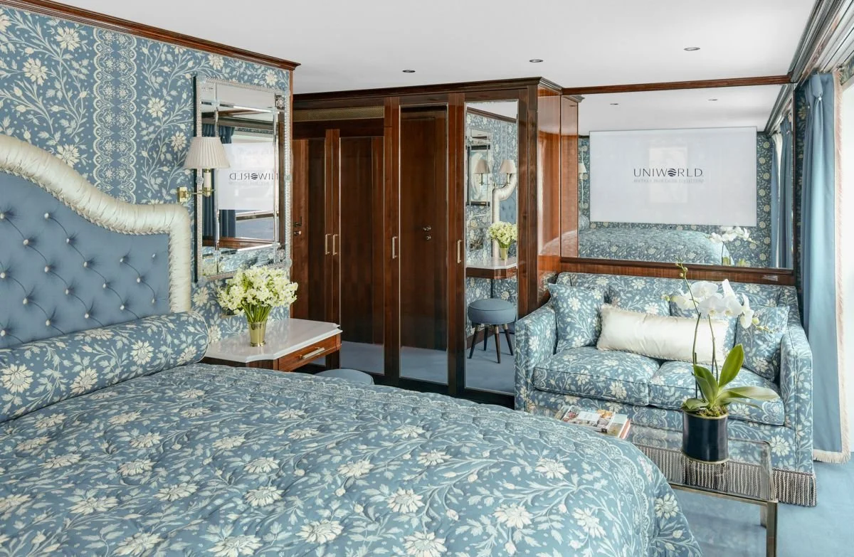Luxurious hotel room with blue floral wallpaper, a bed with a tufted headboard, a side table with a flower vase, a blue sofa with patterned cushions, a small round stool, a mirror, and a projector screen with the word 'UNIWORLD' displayed.