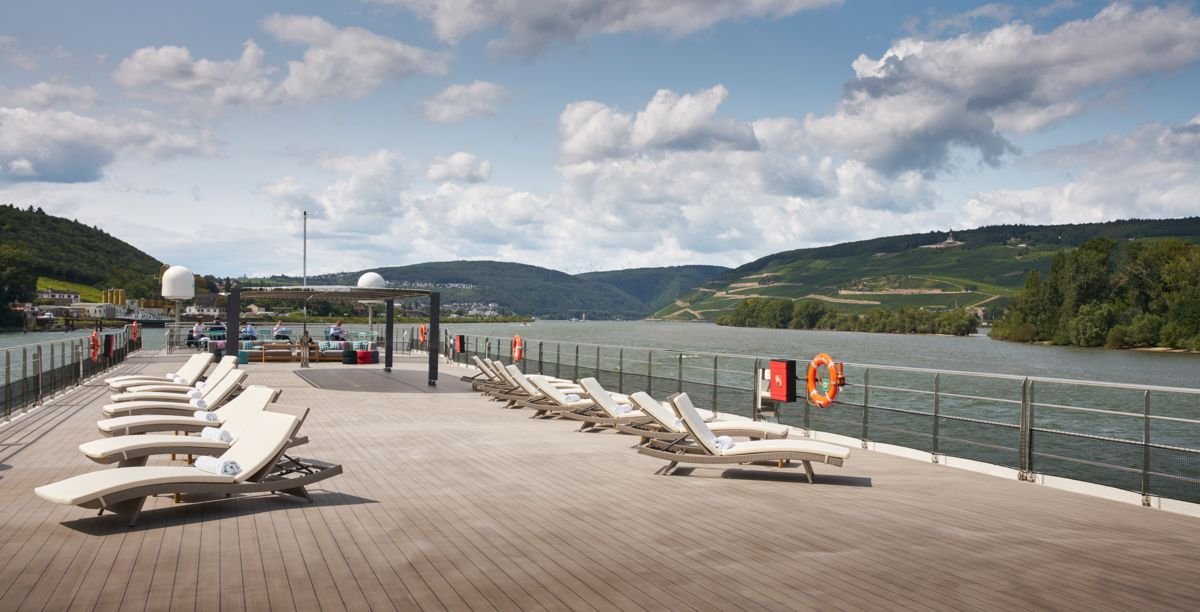 Empty lounge chairs on a cruise ship deck overlooking a river and green hills under a partly cloudy sky.