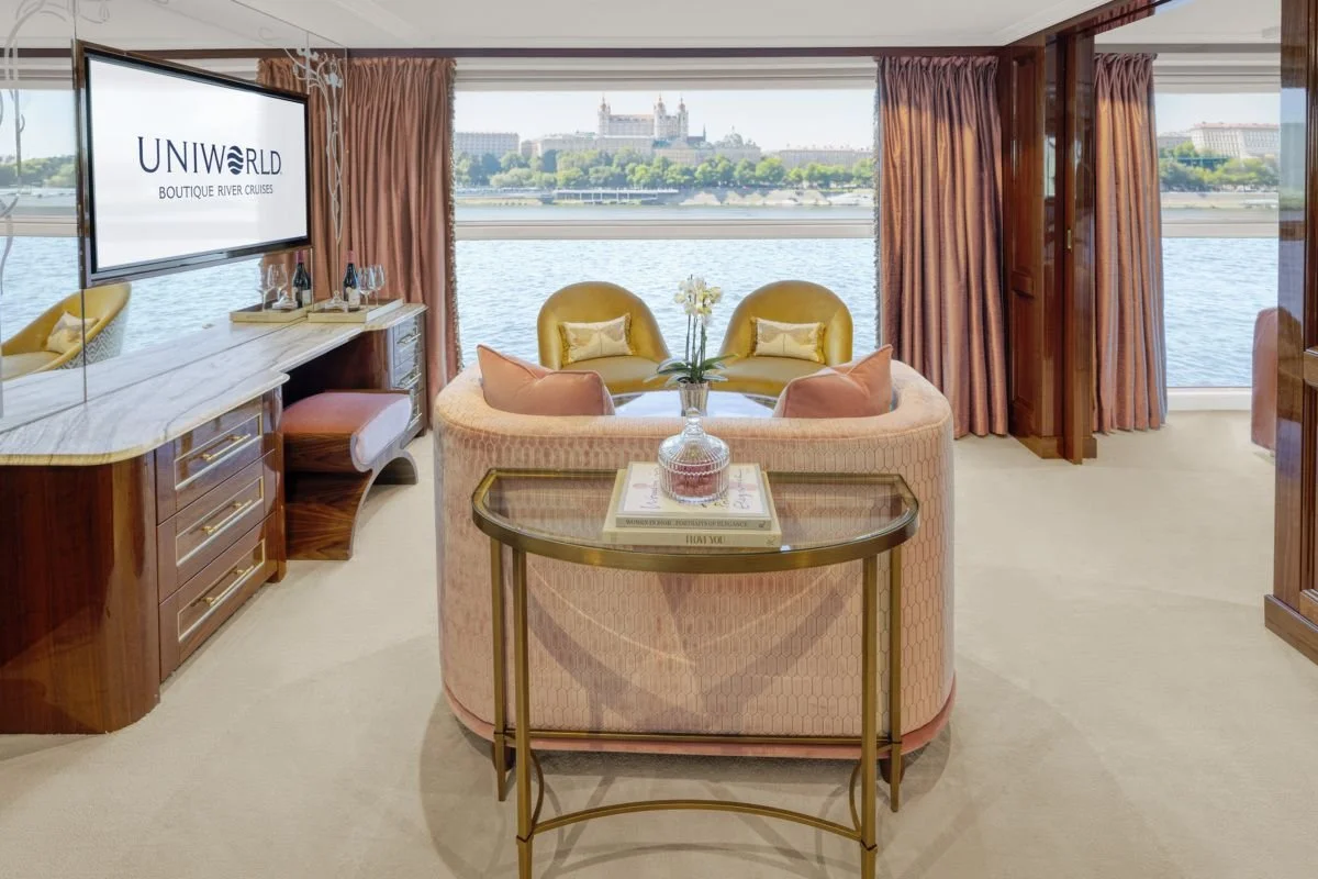 Upscale living room with a large window overlooking a river and a castle in the distance, featuring pink and yellow upscale furniture, a television displaying 'UNIWORLD BOUTIQUE RIVER CRUISES,' and decorative accessories.