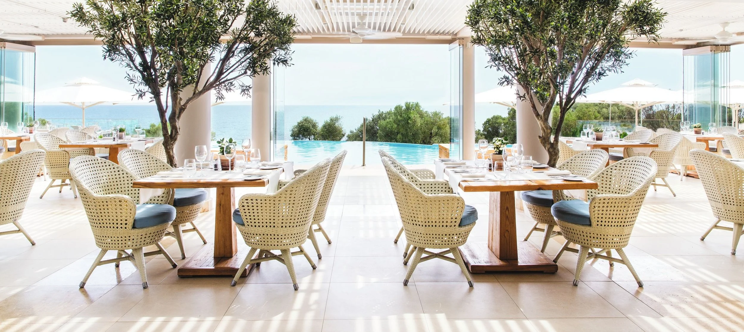 Elegant restaurant with white wicker chairs and wooden tables, outdoor view of the ocean, pool, trees, and umbrellas.