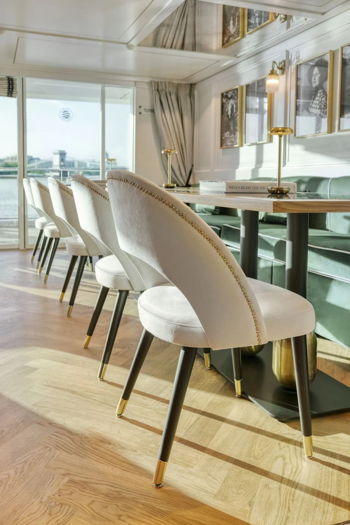 Modern dining area with white upholstered chairs featuring gold accents, a wooden table, green cushioned bench, gold table lamps, black and white artwork on the wall, and sliding glass doors with a city view in the background.
