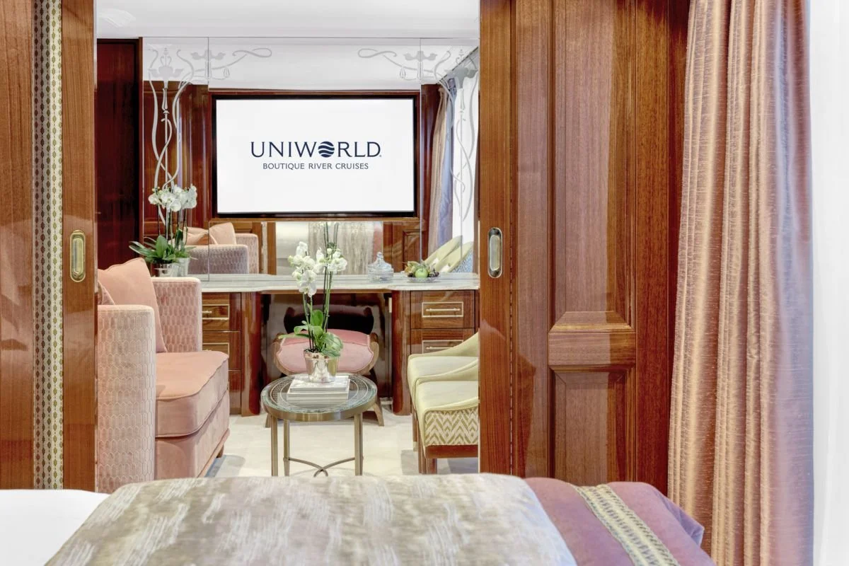 View of a hotel room or suite with a sliding wooden door open to a sitting area featuring pink and cream chairs, a small table with a plant, and a flat-screen TV displaying 'UNIWORLD BOUTIQUE RIVER CRUISES'.