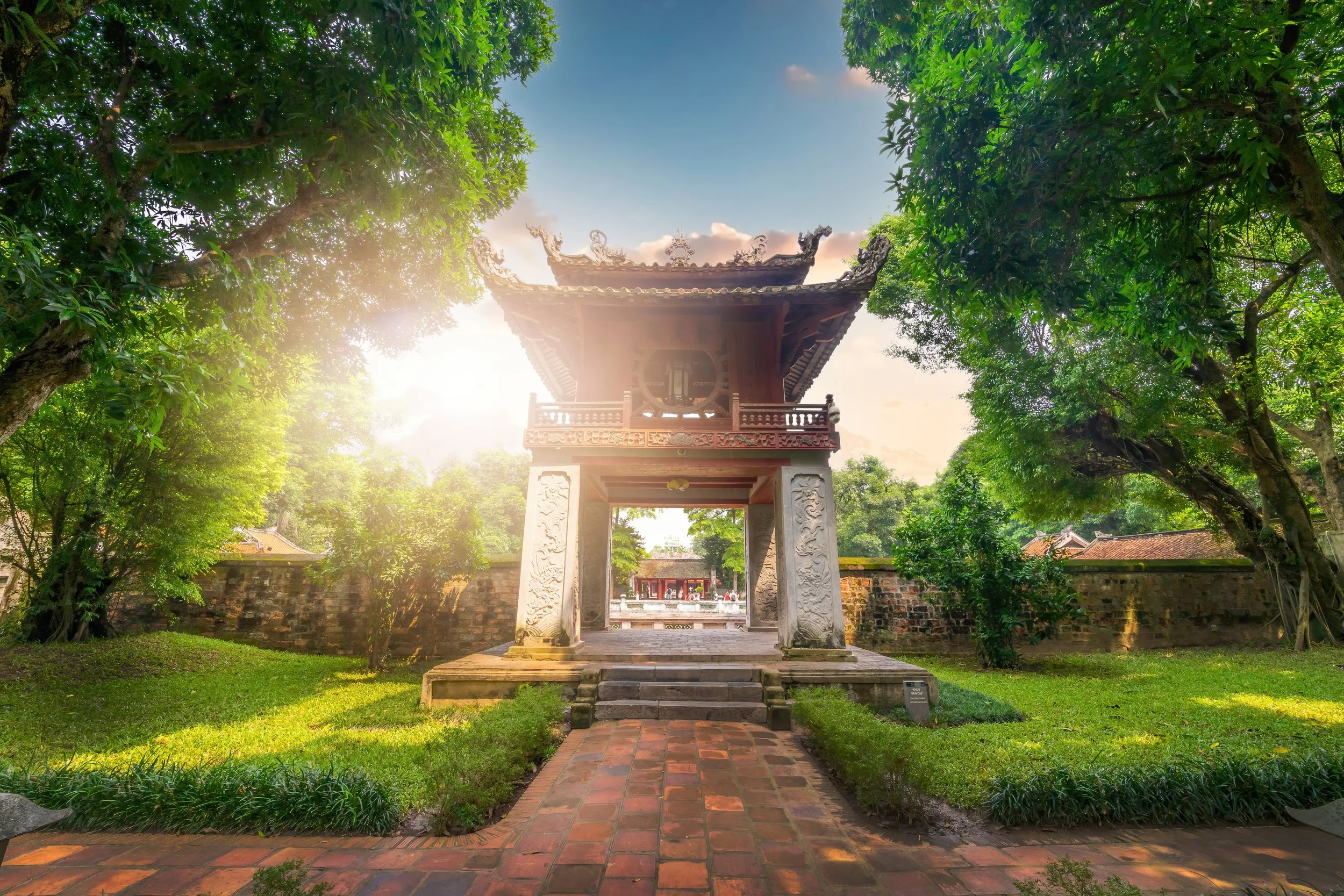A traditional Asian pagoda-style structure with a decorative roof, situated in a lush green park, with sunlight filtering through the trees.