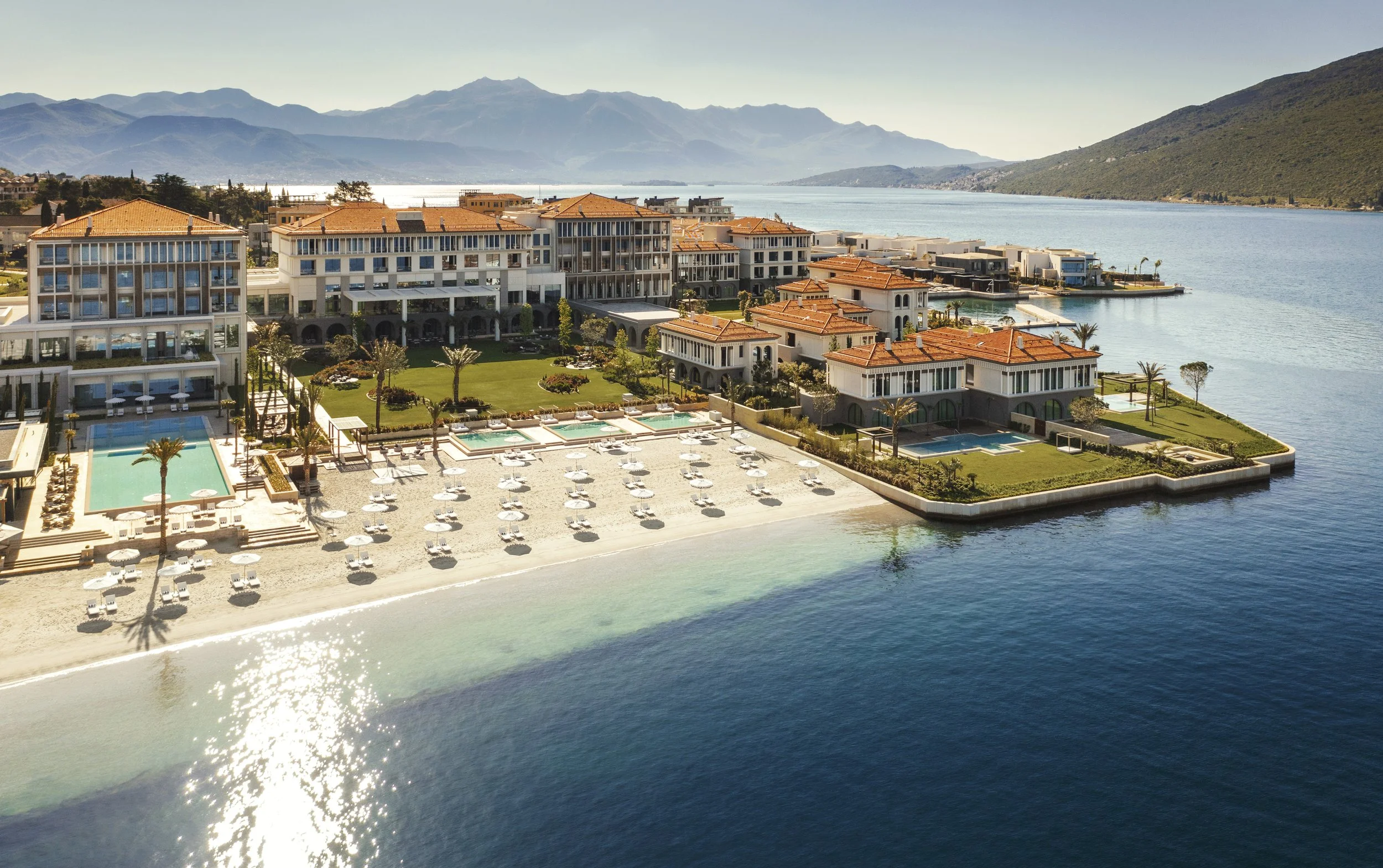 A luxurious waterfront resort with multiple buildings, outdoor pools, palm trees, and sun umbrellas on a sandy beach, set against a backdrop of mountains and a large body of water.