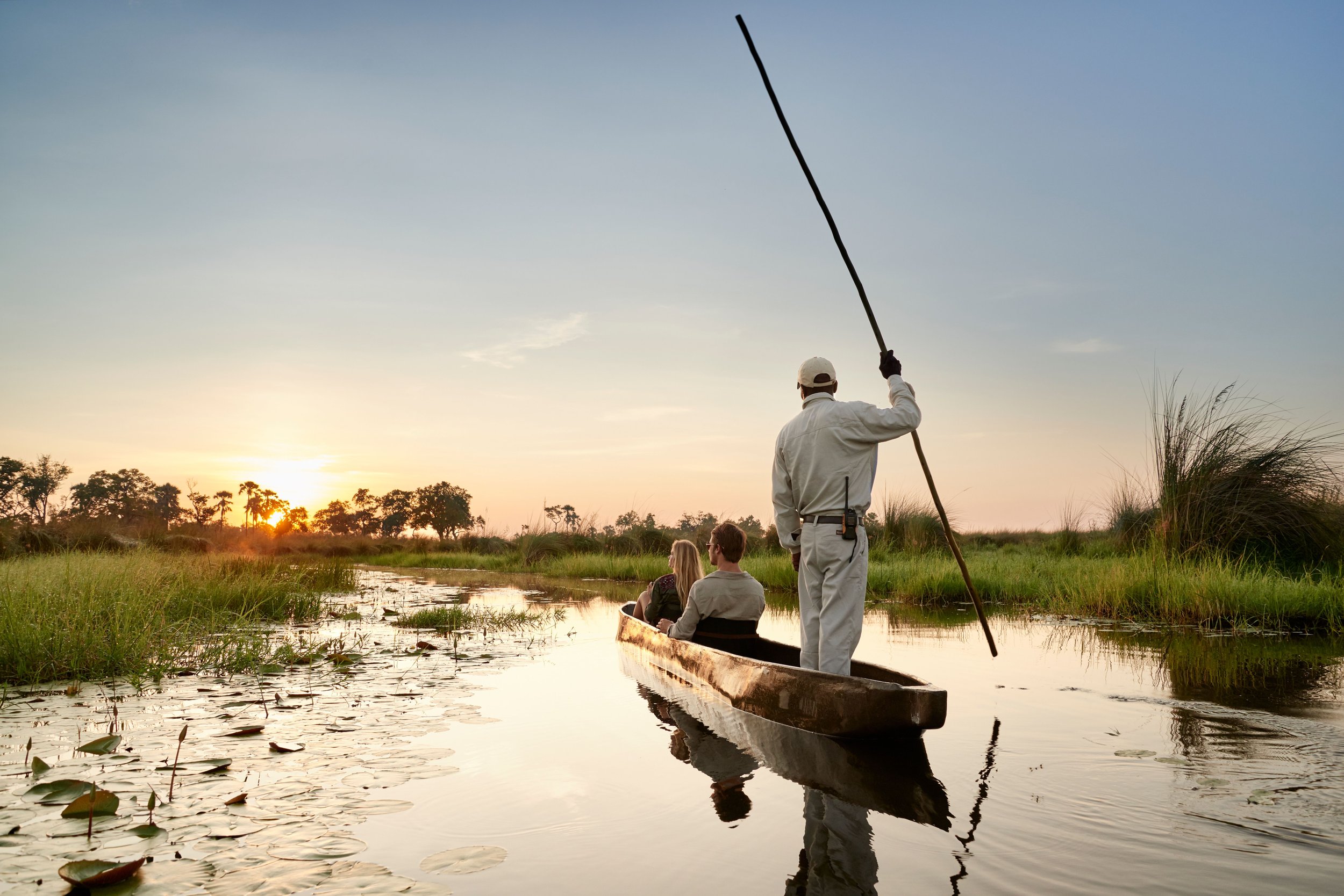 A person standing in a canoe on a river at sunset, holding a pole, with two people sitting at the front of the canoe. The scene is surrounded by tall grass, trees, and water lilies.
