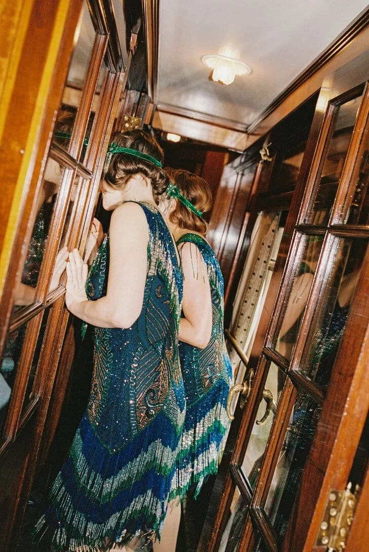 Two women in vintage-style, beaded dresses with feathered hems, wearing green headbands, peering through a small window in a narrow wooden-paneled hallway.