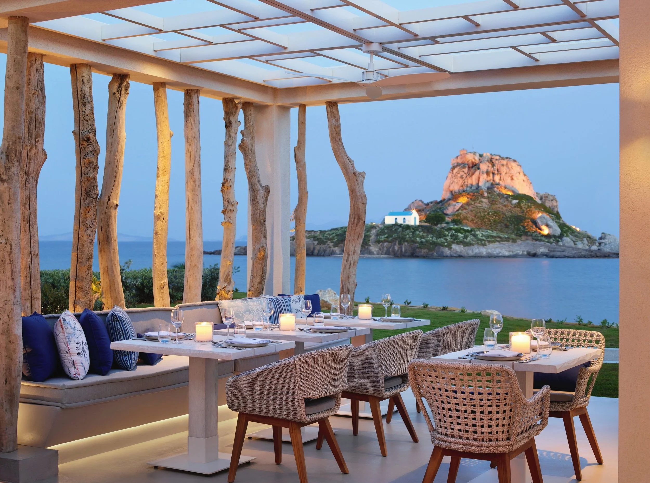 Elegant outdoor dining area with a table set with candles, glasses, and plates, overlooking a coastal landscape with a rocky island and small white chapel visible in the distance during sunset.