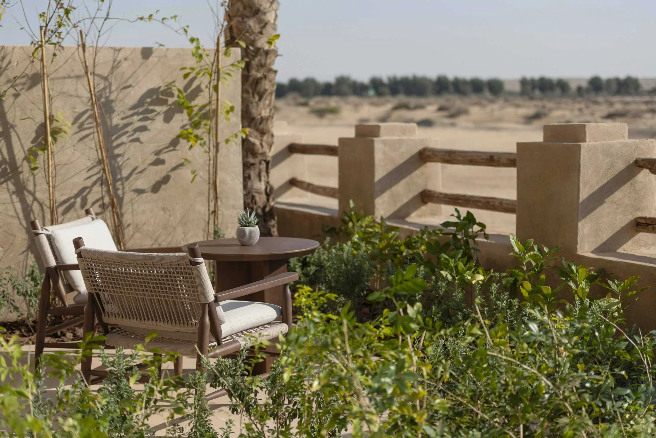 Outdoor patio area with two beige chairs, a small round table with a potted succulent, surrounded by green plants and bushes, with a desert landscape and a concrete wall in the background.