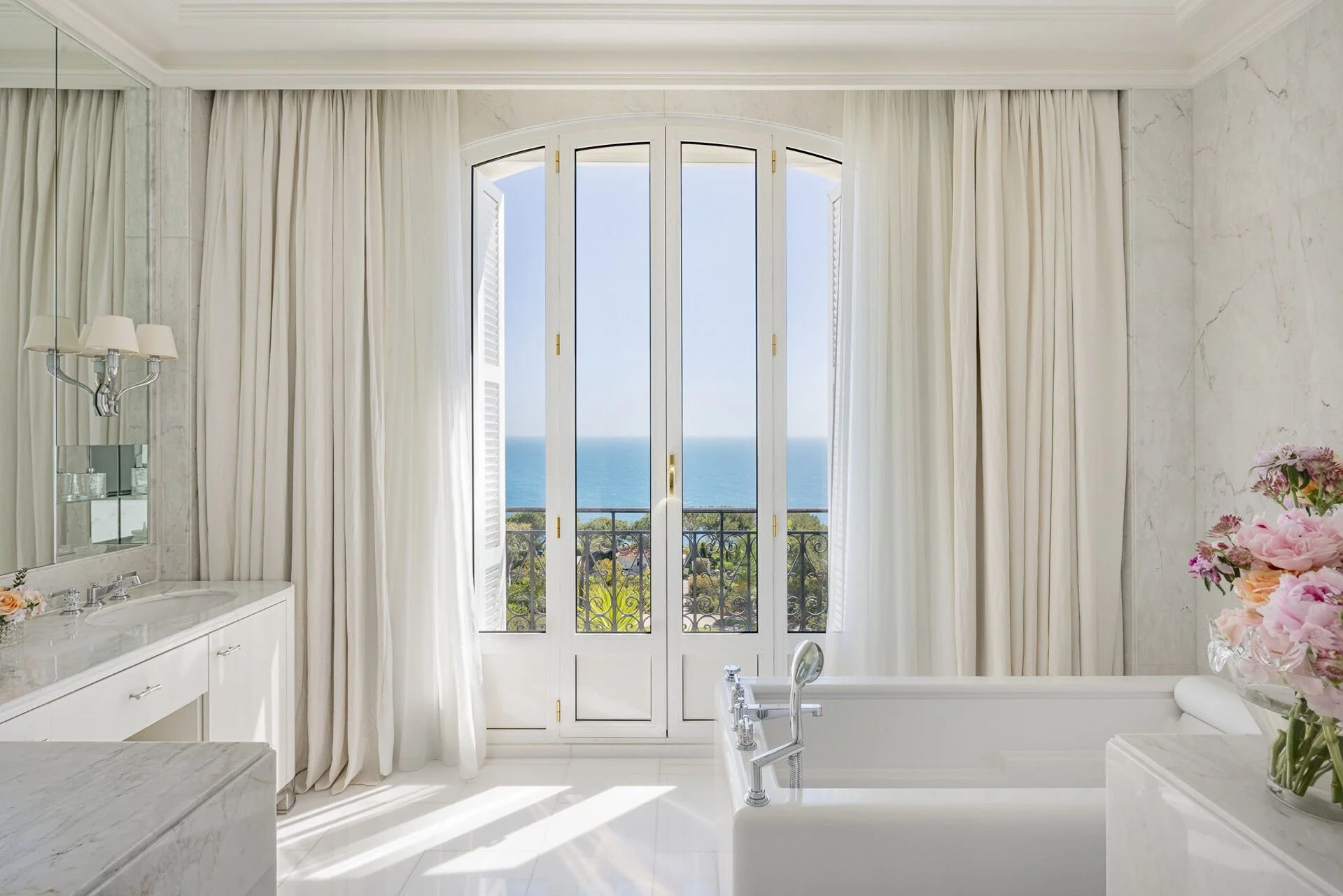 A bright bathroom with a large window overlooking the ocean, with white curtains, a bathtub, a marble vanity with a mirror, and pink flowers on a side table.