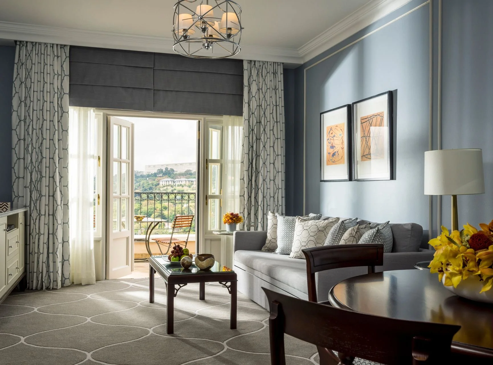 Living room with blue walls, white sofa with patterned pillows, wall art, coffee table with fruit, opened balcony doors with outdoor seating, curtains, chandelier, and a side table with a lamp.