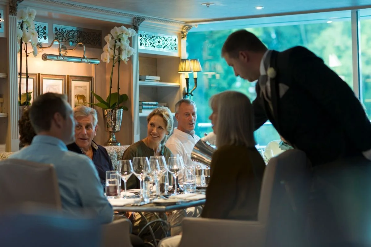 People dining at a formal gathering in a well-decorated restaurant with large windows, flowers, and elegant lighting.
