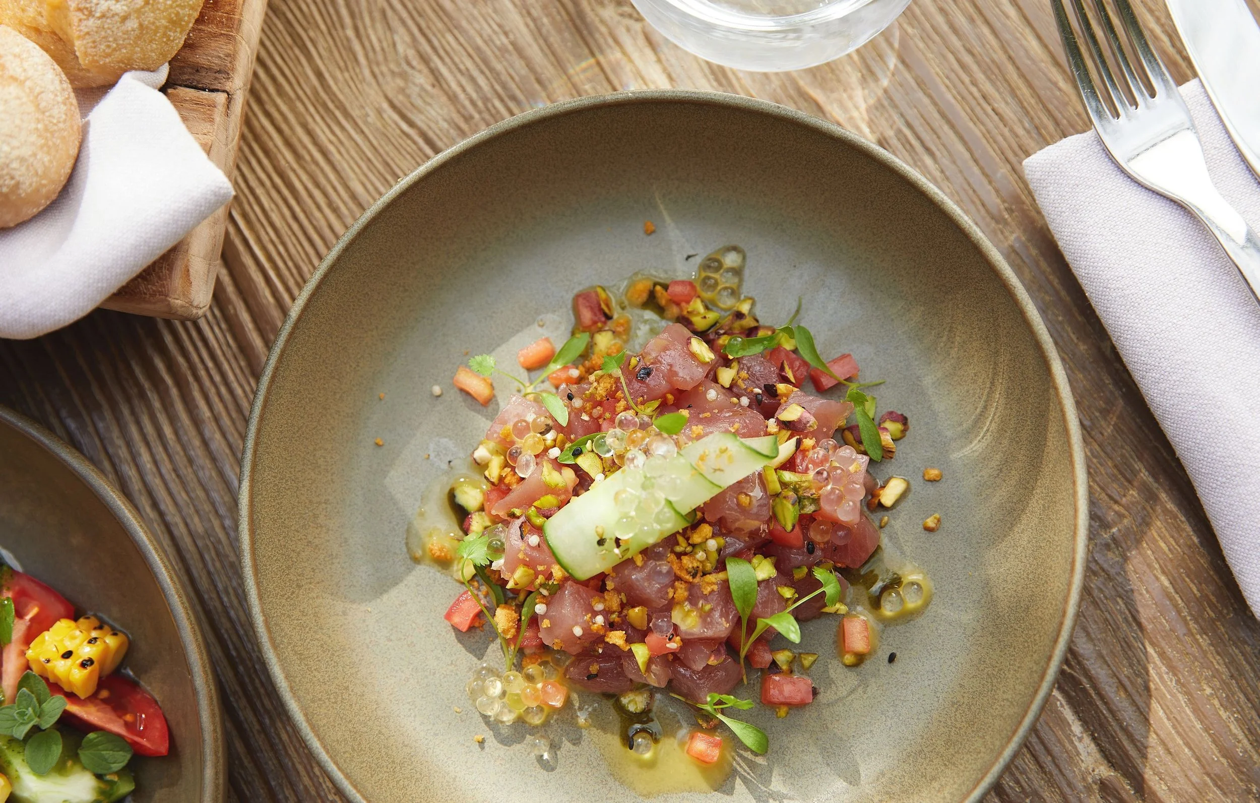 A plate of tuna tartare garnished with chopped pistachios, microgreens, and small spheres of sauce, set on a wooden table with a napkin, fork, and a glass of water nearby.