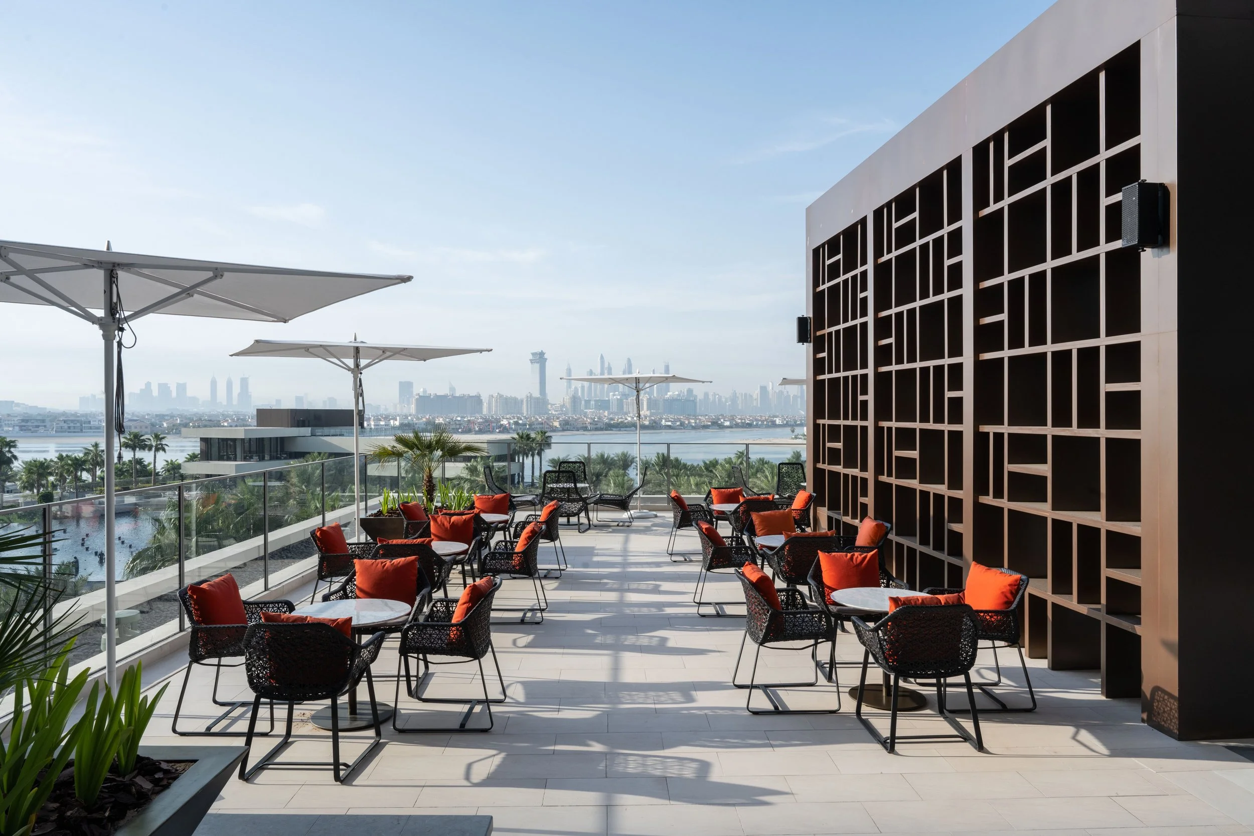 Outdoor rooftop lounge with black chairs and orange cushions, white tables, white umbrellas, overlooking a city skyline with tall buildings and palm trees.