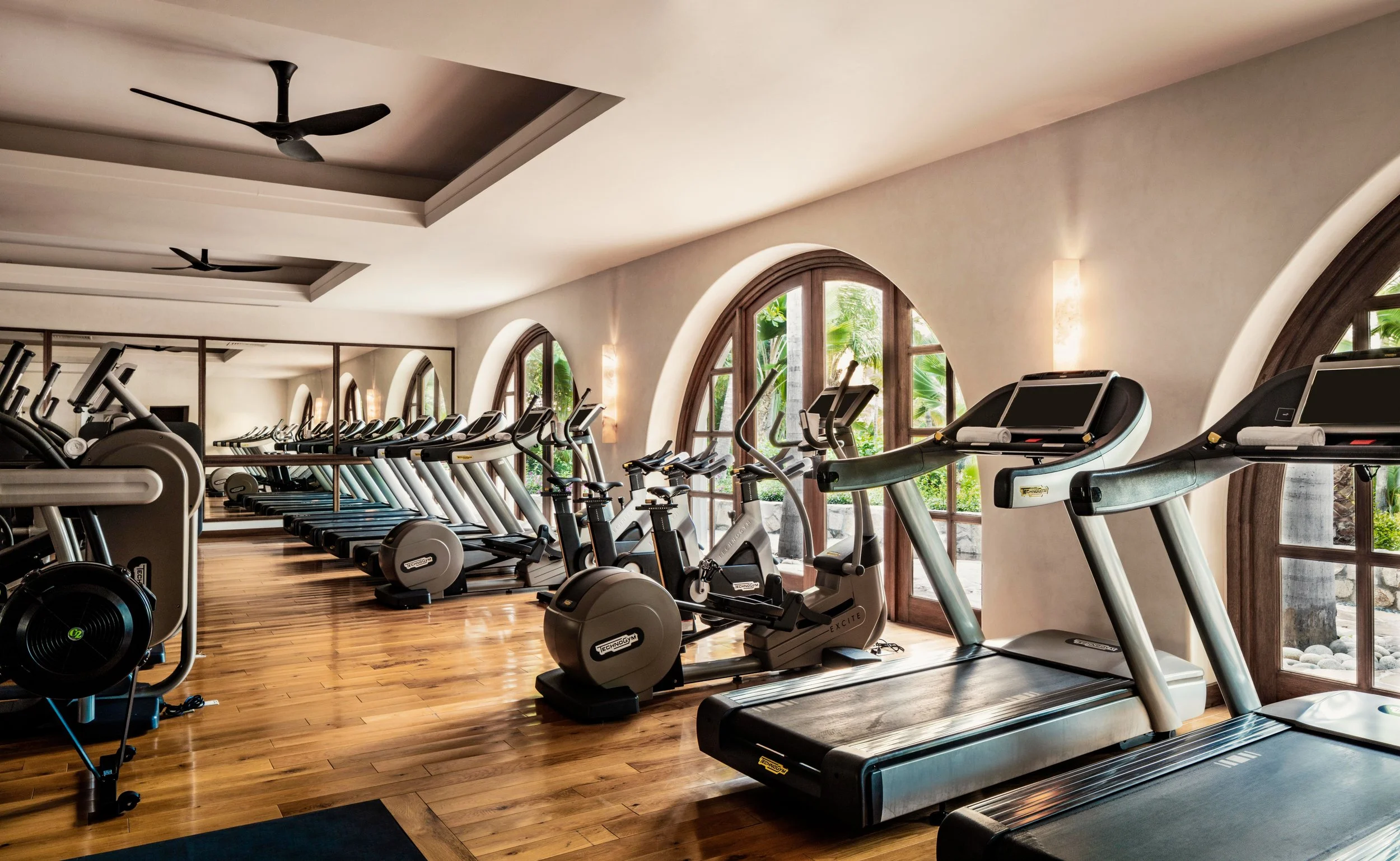 A gym with multiple treadmills and exercise bikes lined up in front of arched windows showing greenery outside, with wooden floors and soft lighting.