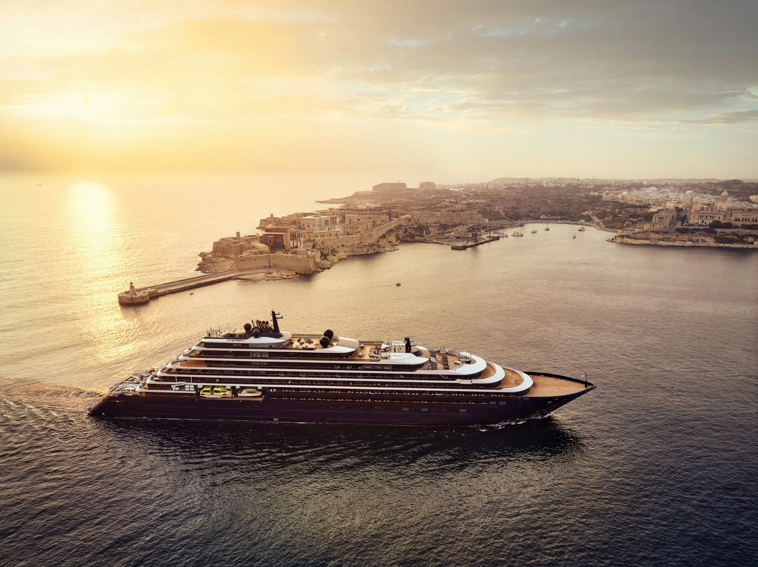 A large luxury yacht sailing in calm waters near a coastal city during sunset.