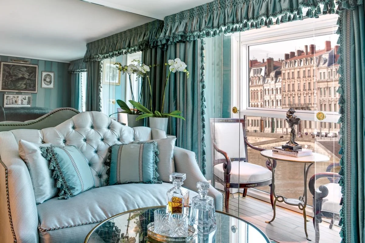 A luxurious living room with a tufted white sofa adorned with coordinating pillows, a glass coffee table with decanters, and a large window with teal curtains. Outside the window, there are historic buildings and a cityscape.