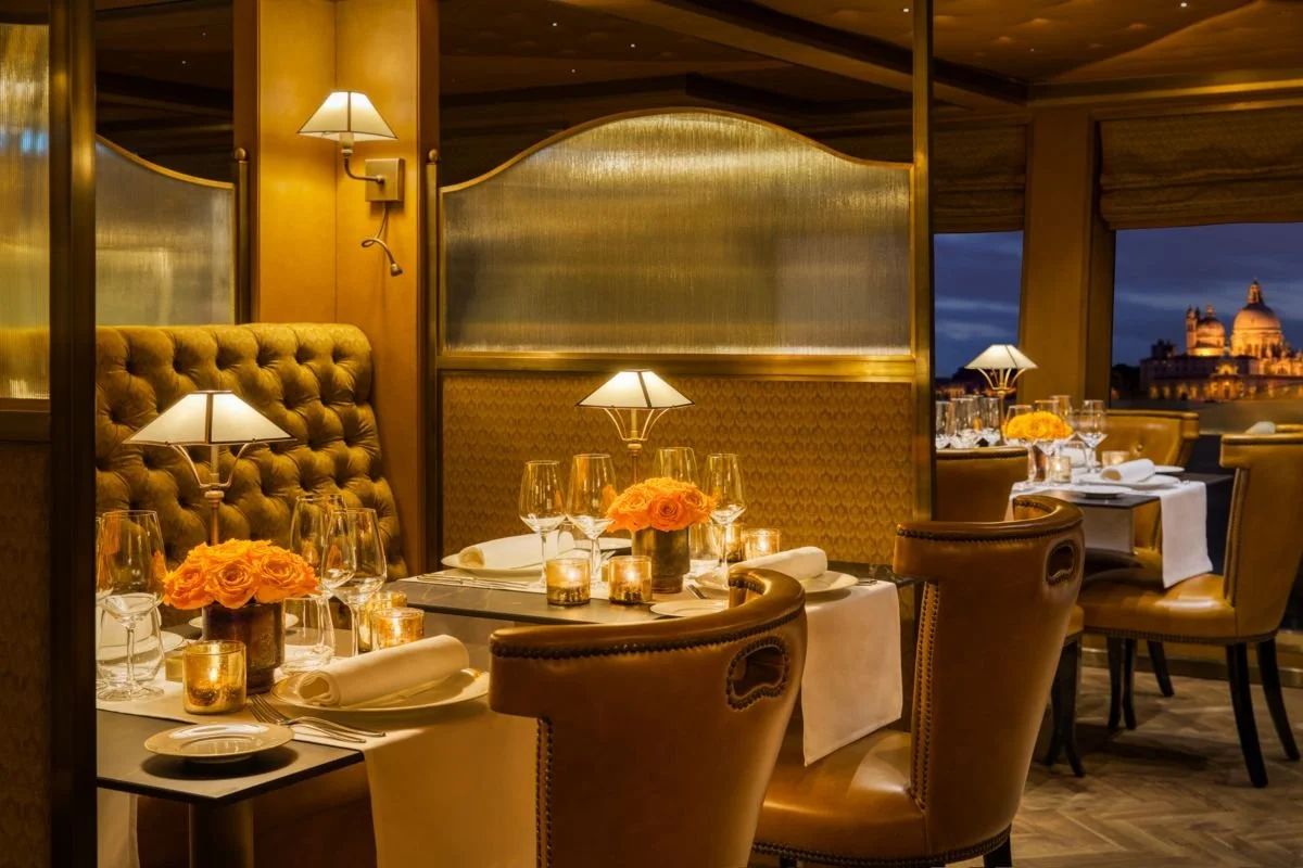 A luxurious restaurant dining room with romantic lighting, set for a formal dinner with tables decorated with peach-colored roses, candles, wine glasses, and folded napkins, overlooking a cityscape at dusk.