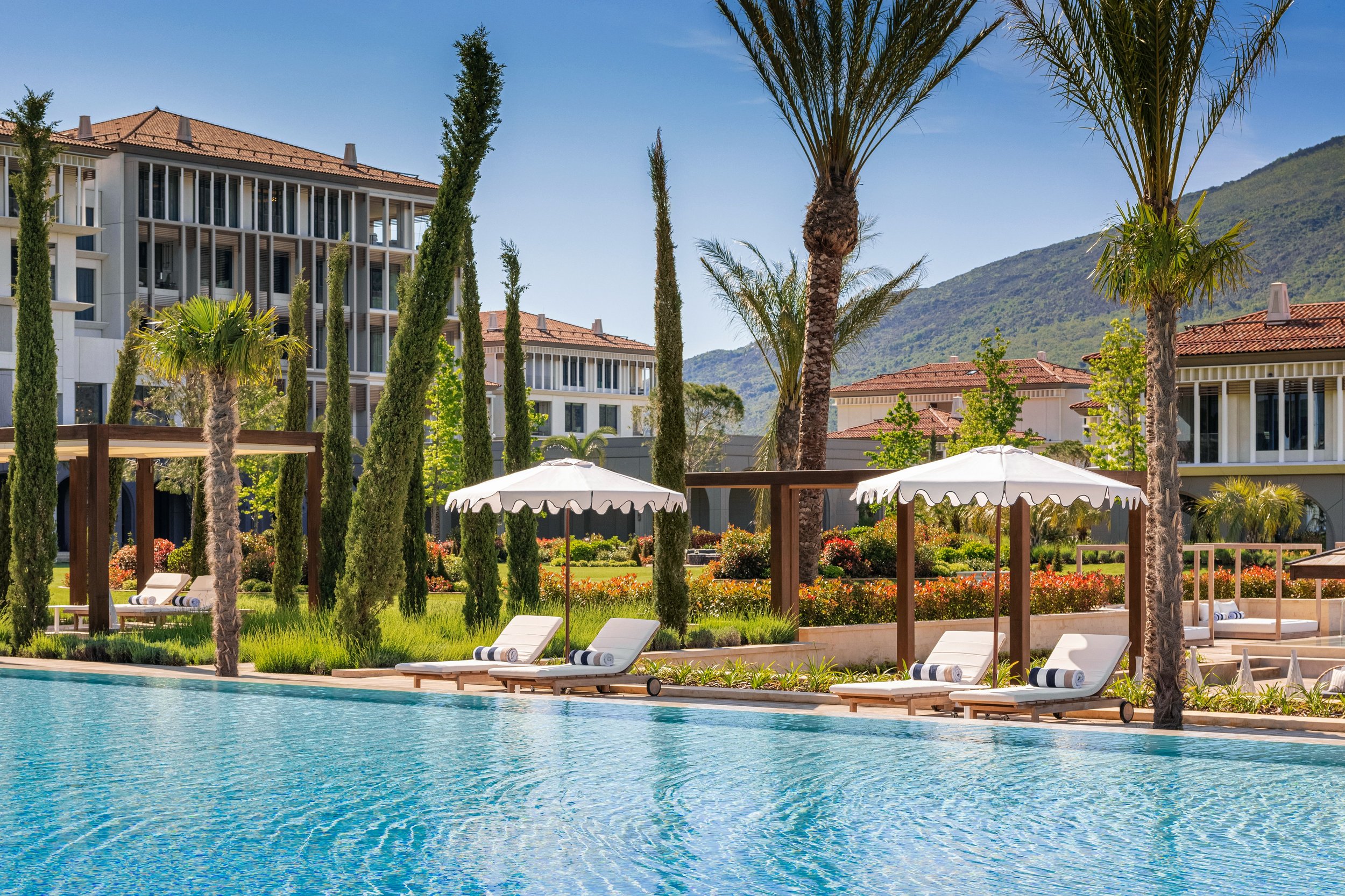 Resort poolside area with lounge chairs, umbrellas, tall palm trees, and landscaped garden with buildings and mountains in the background.