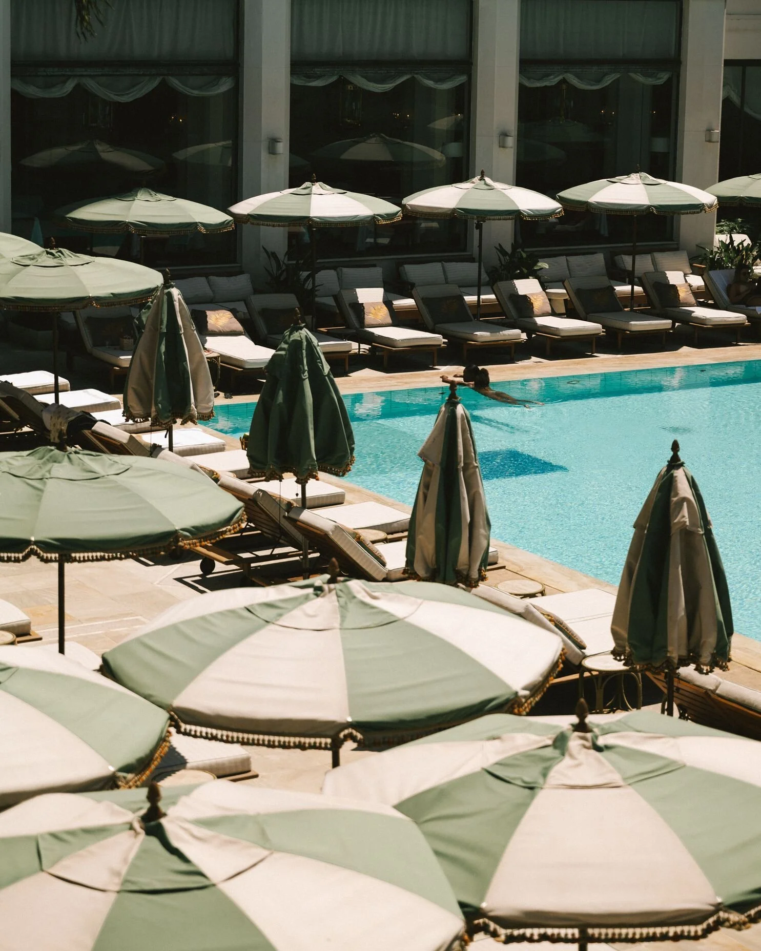A person swimming in a hotel pool with lounge chairs and green umbrellas around the pool.