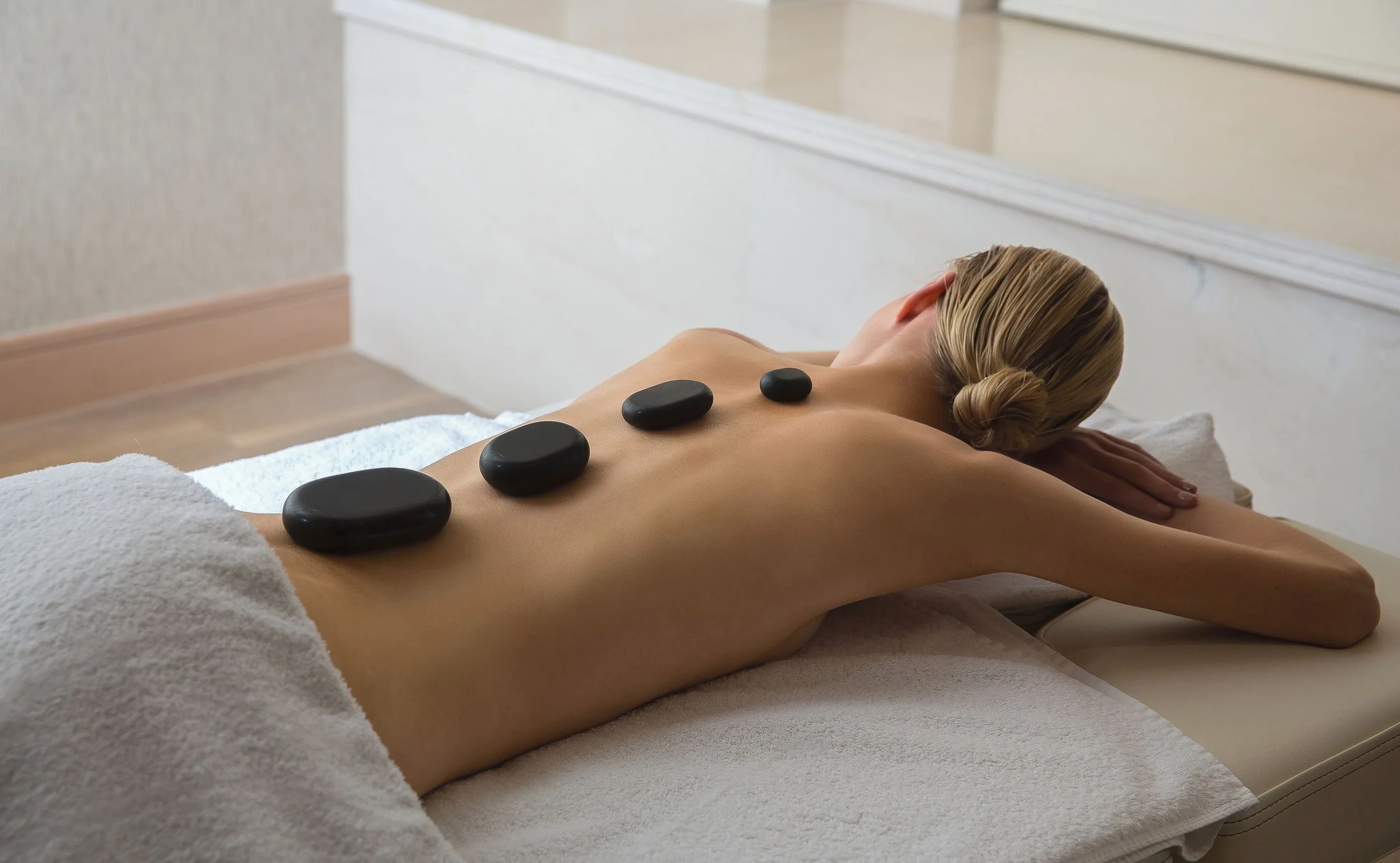 A woman lying face down on a massage table with four black heated stones placed along her back during a massage treatment at a spa or wellness center.