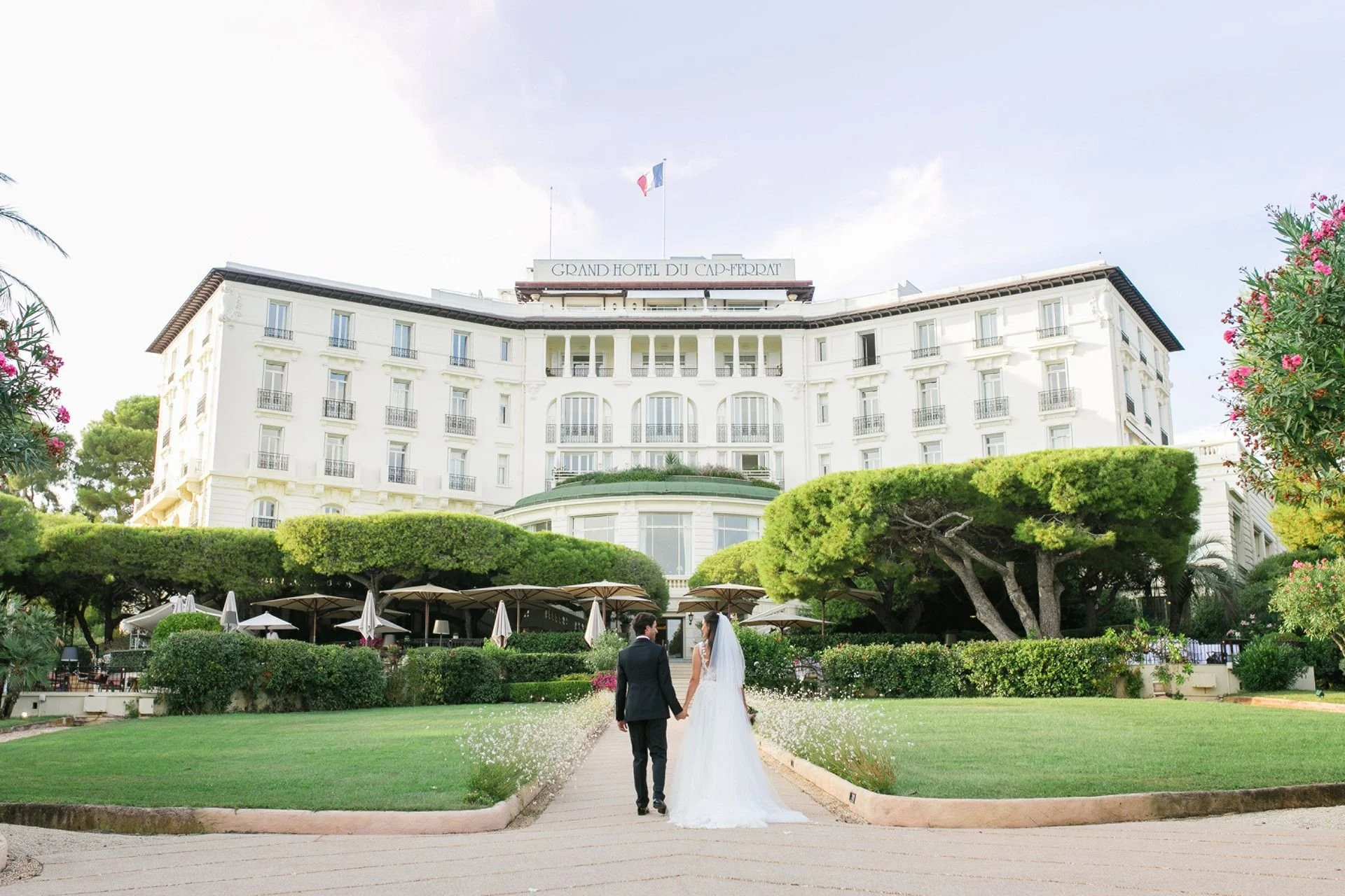 A bride and groom holding hands walking toward a grand hotel with white facade, multiple windows, and a sign reading 'Grand Hotel du Cap-Ferrat,' surrounded by lush green trees and flowering plants.