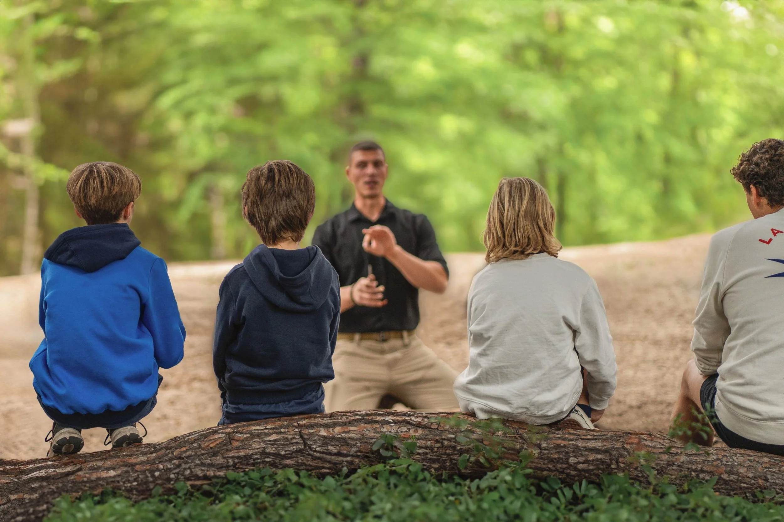 A group of children sitting on a fallen log in a forest, listening to a man giving a talk or presentation.