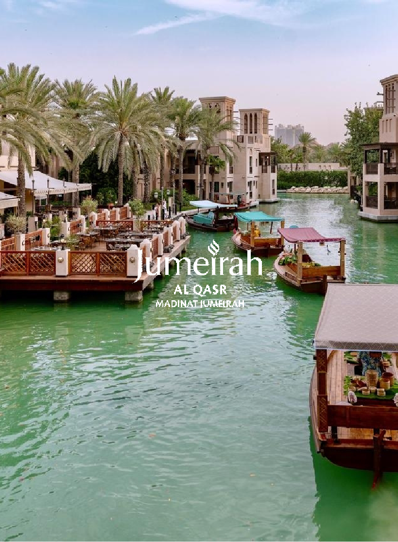 View of a man-made waterway lined with palm trees, traditional Middle Eastern style buildings, and boats with canopies floating on the water in Jumeirah Al Qasr, Madinat Jumeirah in Dubai.