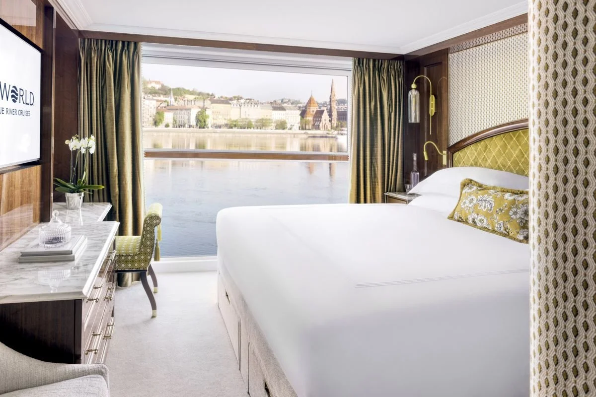 A hotel room with a large window overlooking a river and cityscape, a king-sized bed with green and yellow pillows, a marble-topped desk with a chair, and wall-mounted lamps.