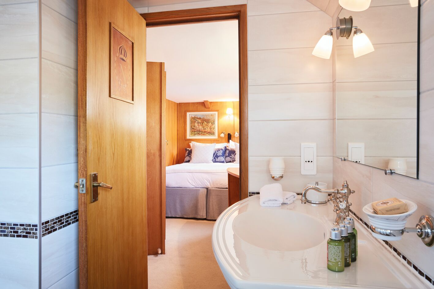 Bathroom with a sink, mirror, wall-mounted lighting, and towels, with an open door leading to a bedroom with a bed, pillows, a painting, and bedside lamps.