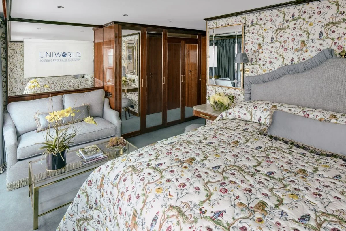 A hotel room featuring a large bed with a floral-patterned bedspread, a gray upholstered headboard, and matching pillows. There's a side table with a floral arrangement. Opposite the bed, a small sitting area includes a gray sofa adorned with a flower pillow, a glass coffee table with books and a plant, and a mirror on the wall. A wooden wardrobe and a small vanity with a stool are also visible.