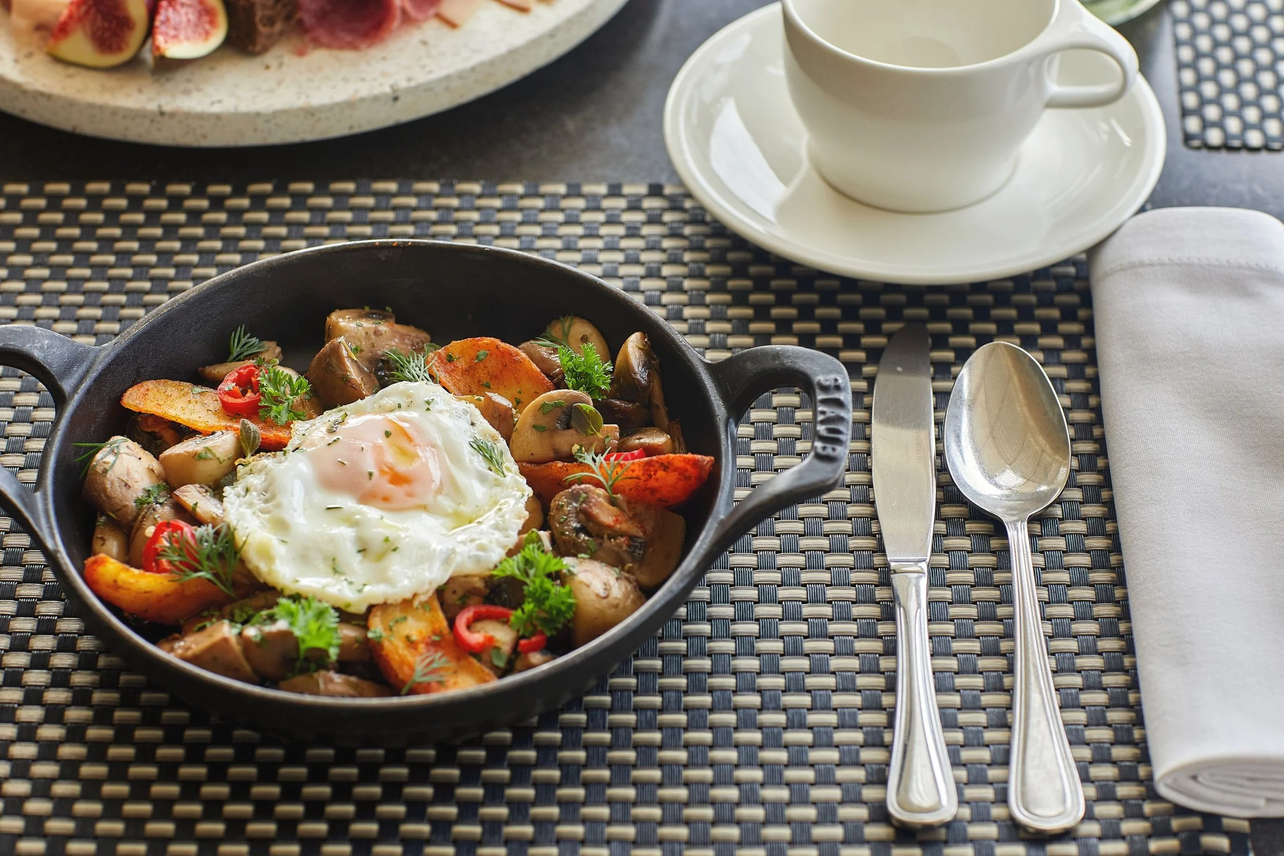 A black skillet with a fried egg on top of sautéed mushrooms, potatoes, and vegetables, garnished with herbs. Nearby are a set of silverware, a white napkin, a white teacup on a matching saucer, and a plate with sliced figs and meat, all on a woven placemat.