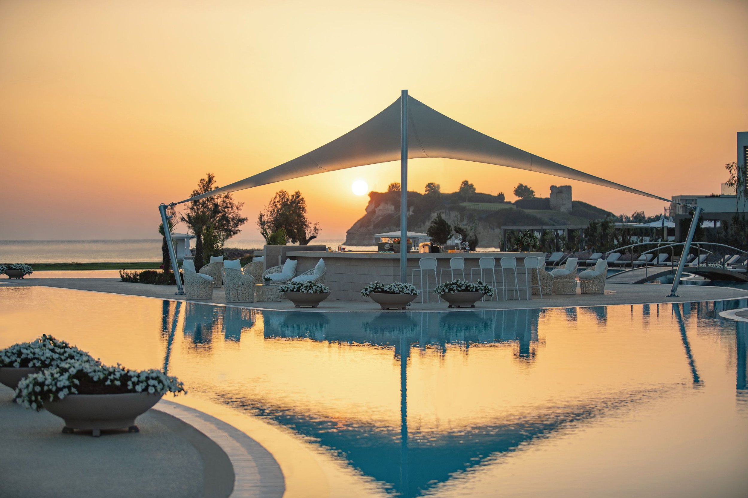 Sunset over a poolside bar area with lounge chairs, umbrellas, and potted flowers, overlooking a rocky coastline with cliffs and a tower in the distance.