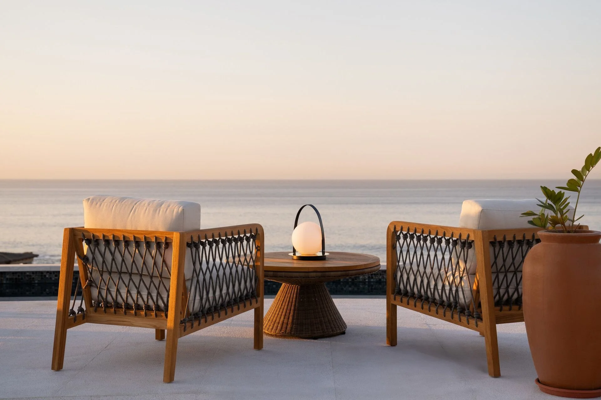Outdoor seating with two wooden chairs, a small round table with a lamp, a potted plant, overlooking the ocean at sunset.
