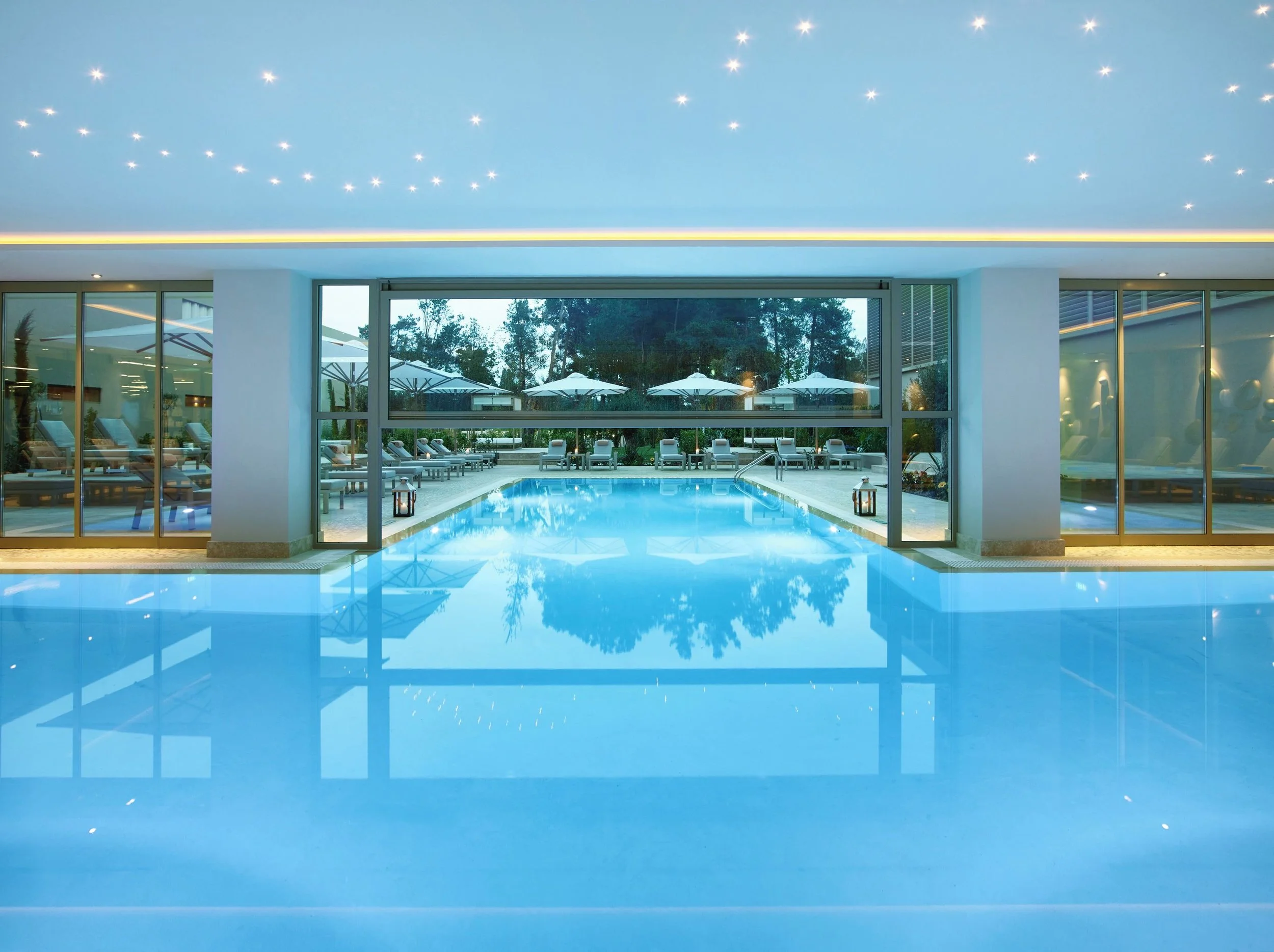 Indoor swimming pool with lounge chairs and umbrellas outside, viewed through large glass doors, under a ceiling with star-shaped lights.