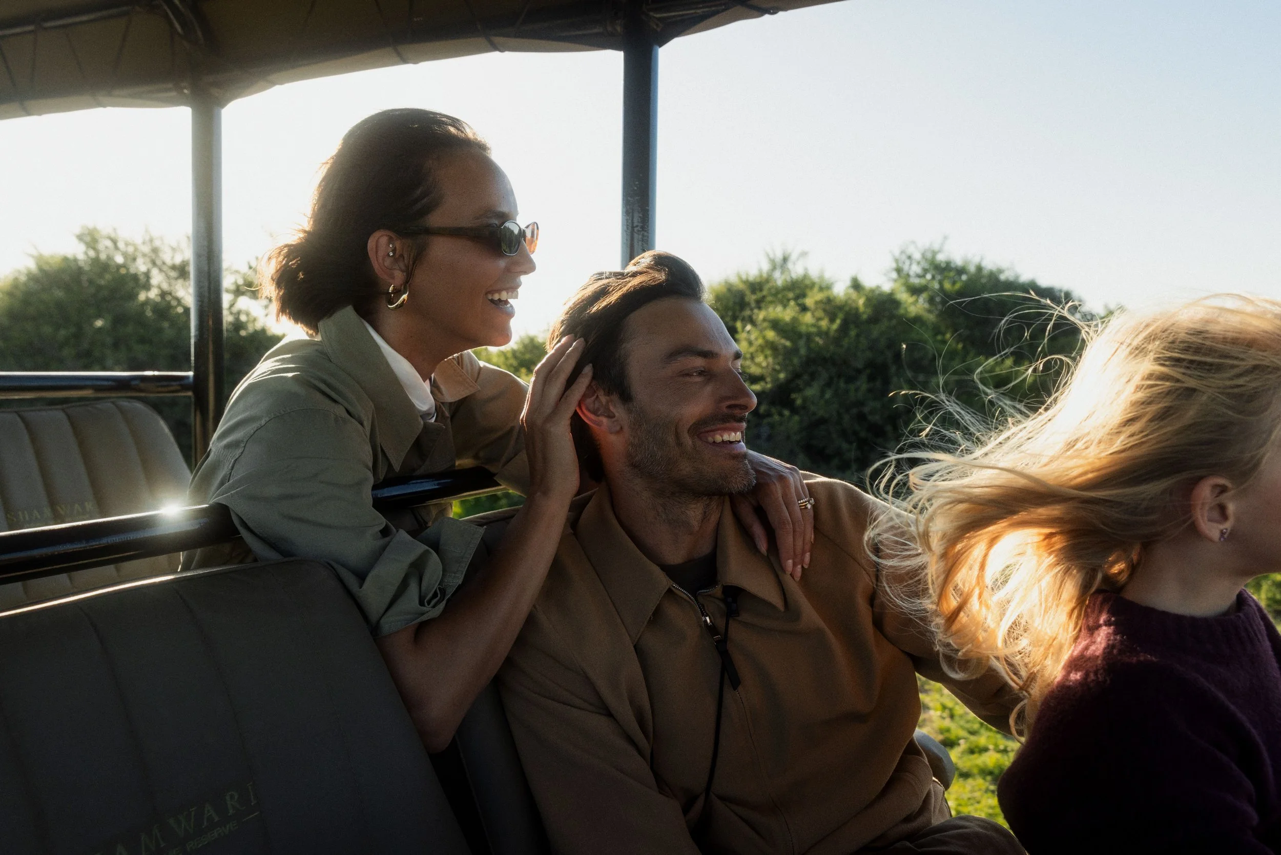 A woman with short hair, sunglasses, and hoop earrings is smiling and leaning on a man with dark hair and a beard, both sitting on a tour bus. Other passengers are visible, and green trees are in the background.