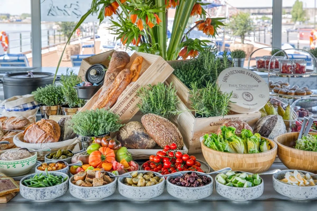A vibrant breakfast spread featuring fresh vegetables, bread, grapes, cherry tomatoes, and herbs displayed on a table with a view of a river outside.