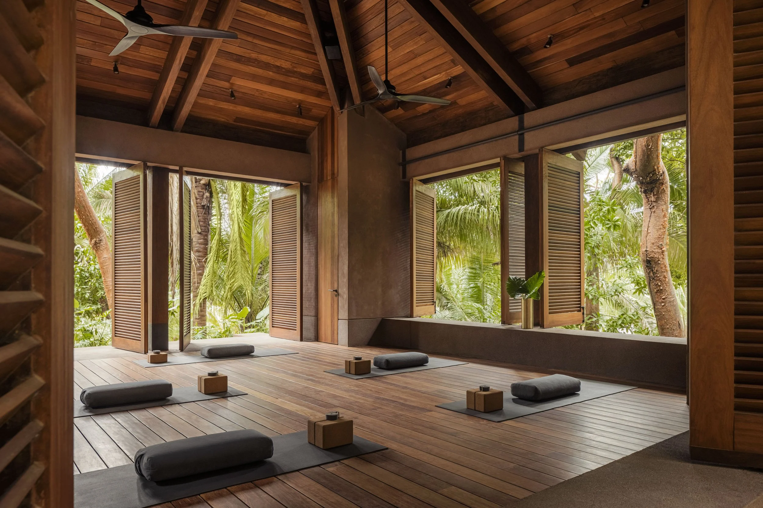 Indoor yoga studio with wooden floors and ceiling, open doors revealing lush tropical trees outside, yoga mats with blocks and bolsters, ceiling fans, and track lighting.