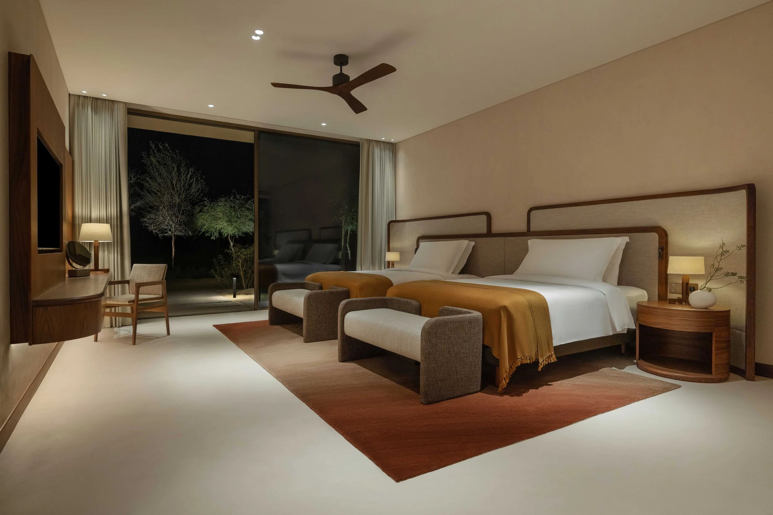 Modern hotel bedroom with two beds, nightstands with lamps, a sitting bench at foot of beds, a desk with chair, a large window with curtains showing trees outside, ceiling fan, and recessed ceiling lights.