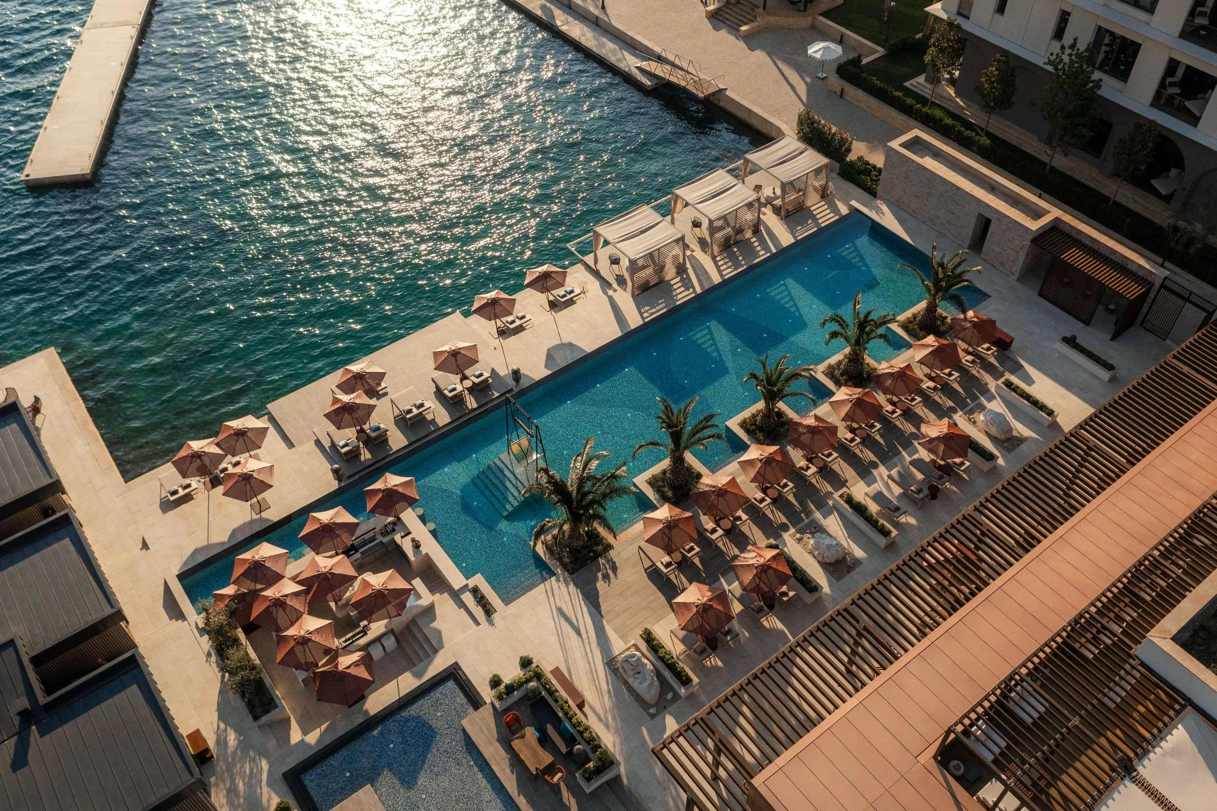 An aerial view of a modern outdoor pool area with lounge chairs, umbrellas, palm trees, and cabanas, adjacent to a body of water, possibly a river or lake, in an urban setting.