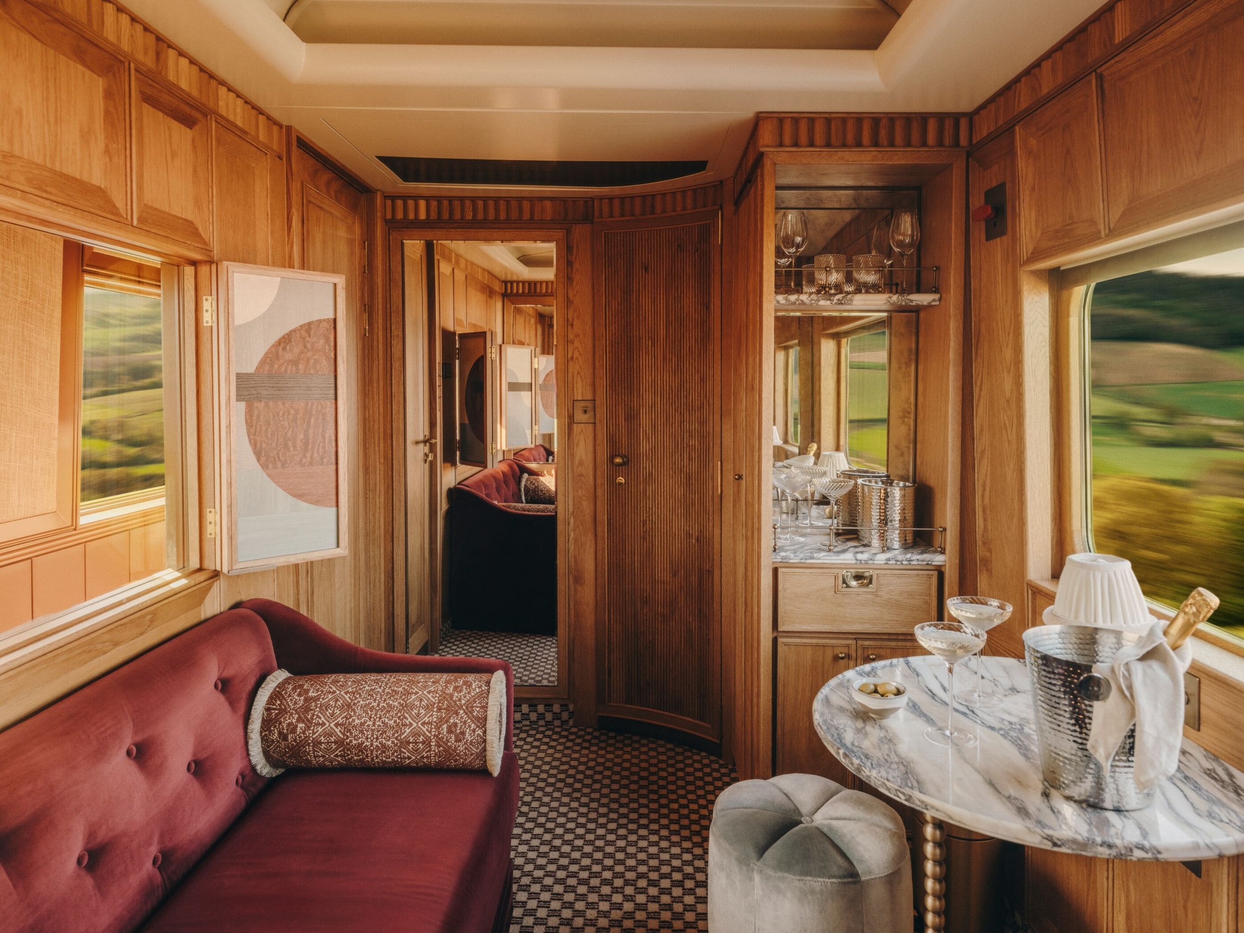 A cozy vintage train living room with wooden paneling, a red tufted sofa with patterned pillow, marble-top round table with glassware, and large windows showing blurred green landscape outside.