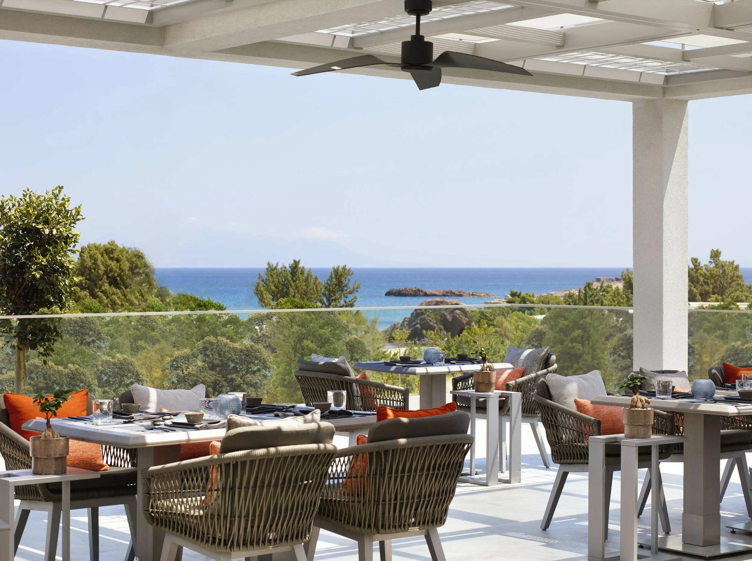 Outdoor dining area with tables set with dishes, glasses, and small vases, under a gazebo with a ceiling fan, overlooking lush greenery and the ocean in the background.
