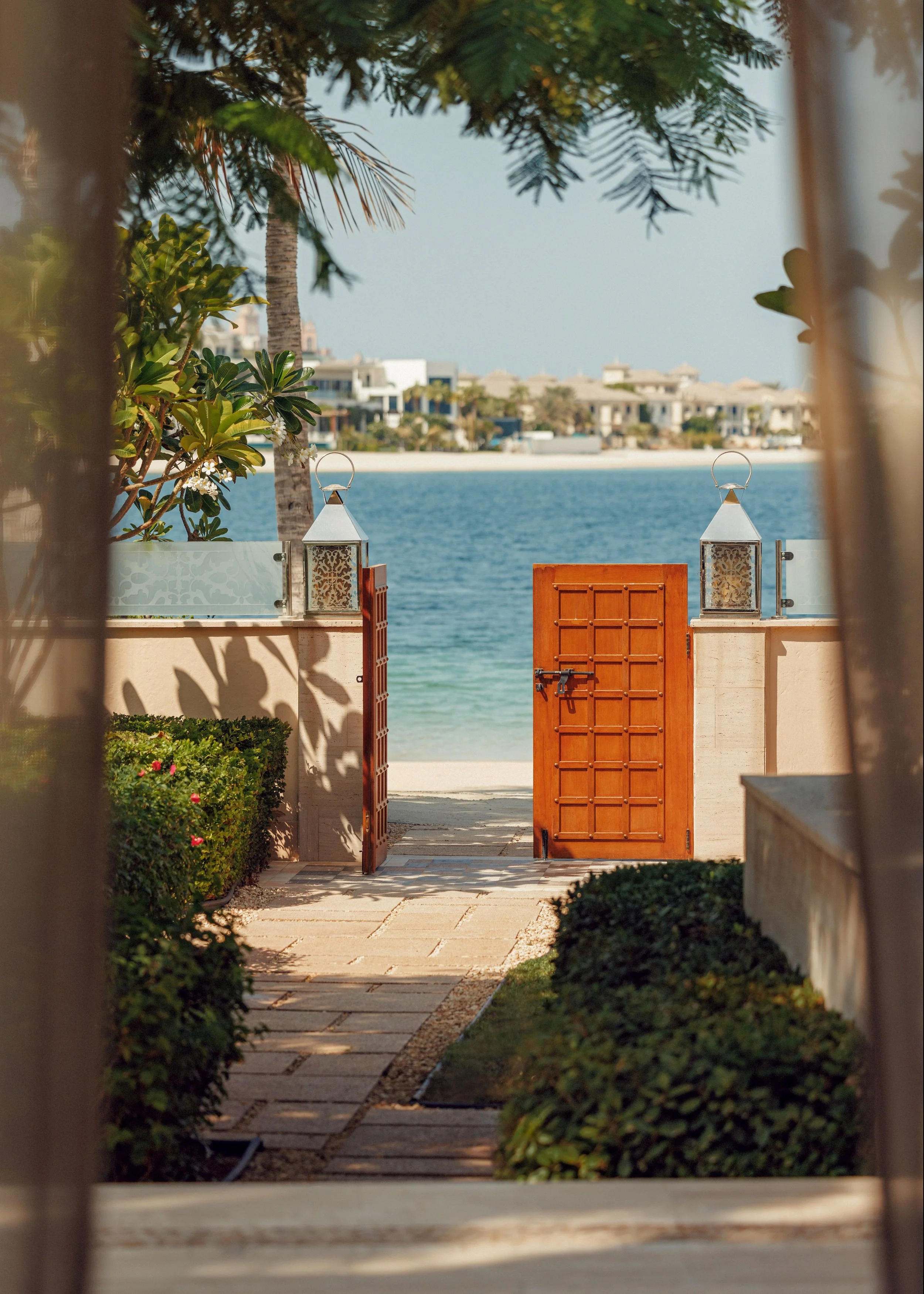 A view through a framed opening of a pathway leading to a wooden gate opening to a beach with water and houses in the background. There are lanterns on each side of the gate, green bushes along the pathway, and trees overhead.