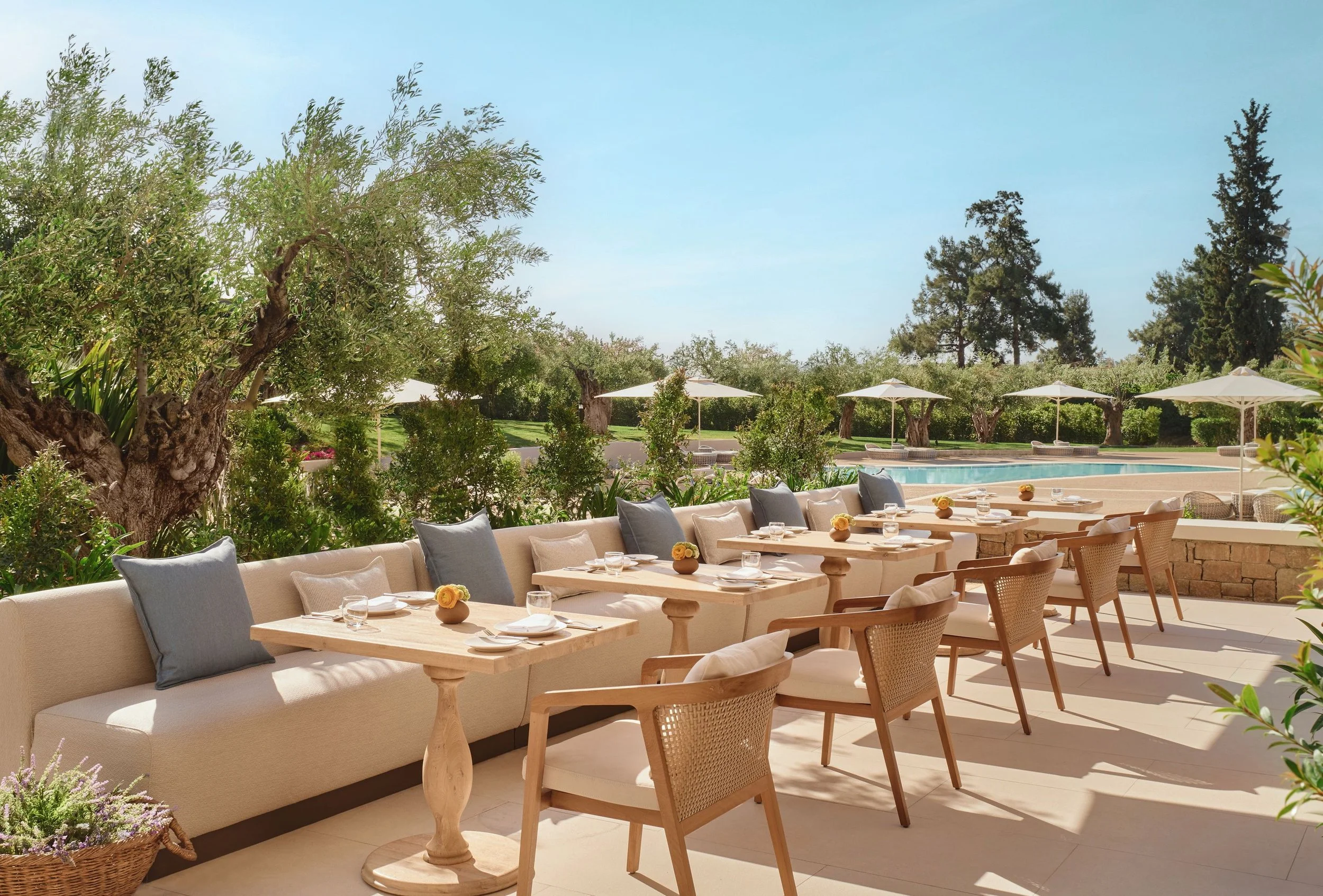 Outdoor dining area with a long sofa with cushions, tables set with glasses and small flowers, surrounded by greenery, trees, pool, umbrellas, and a clear blue sky.