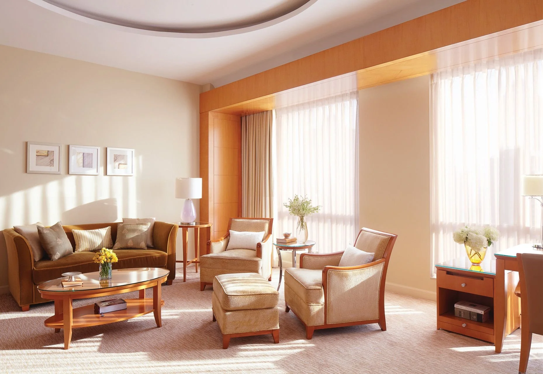 Living room with beige walls, large windows with sheer curtains, a brown sofa with pillows, a set of beige armchairs, glass-top coffee table with flowers, a side table with a lamp, and a wooden entertainment stand.